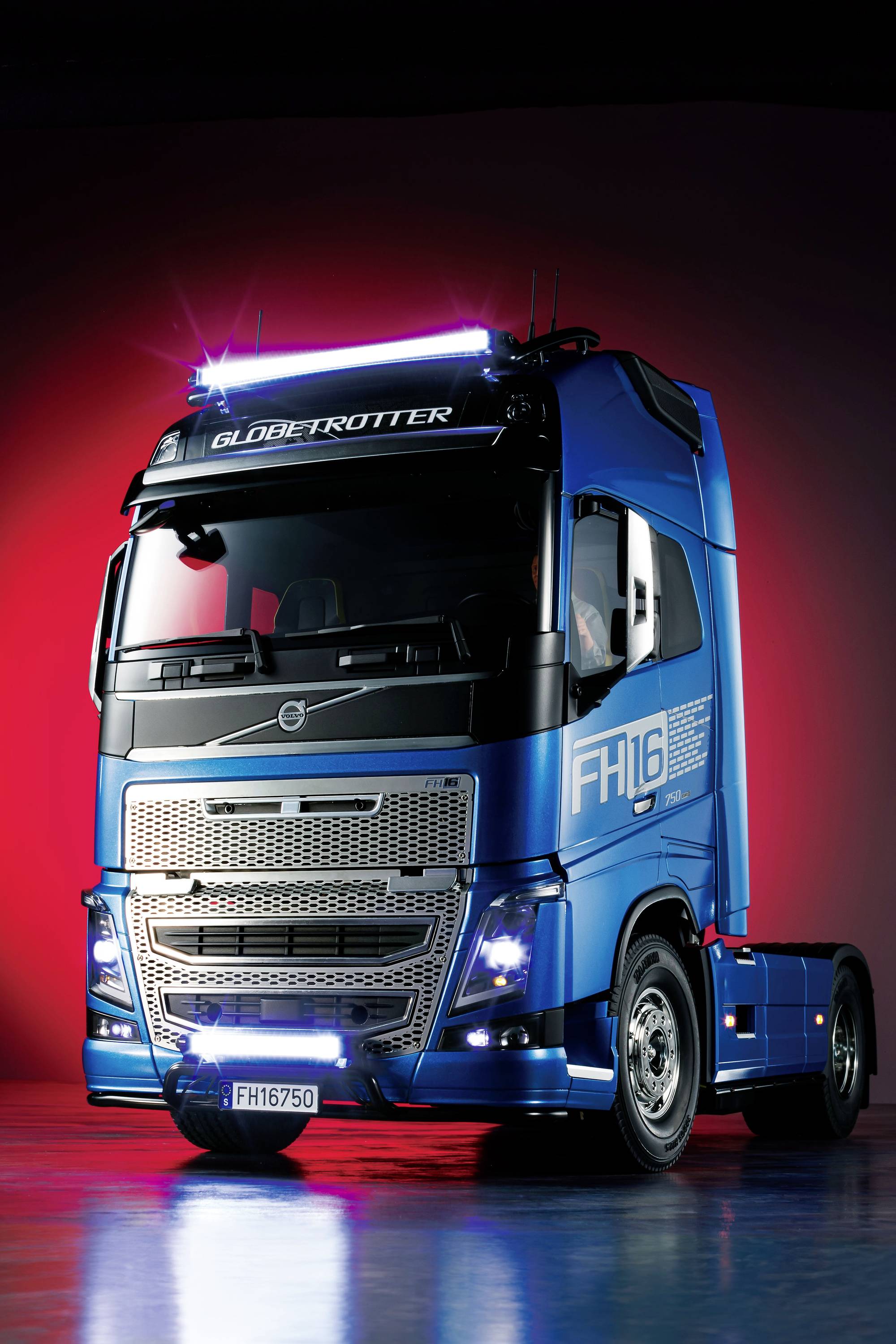 Un camion bleu portant l'inscription 'FH16' sur le côté et 'Globetrotter' au-dessus du pare-brise, éclairé par des lumières rouges.