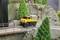 Un camion-miniature jaune roule sur une route miniature dans un paysage montagneux. Des arbres et un piéton sont visibles en arrière-plan.