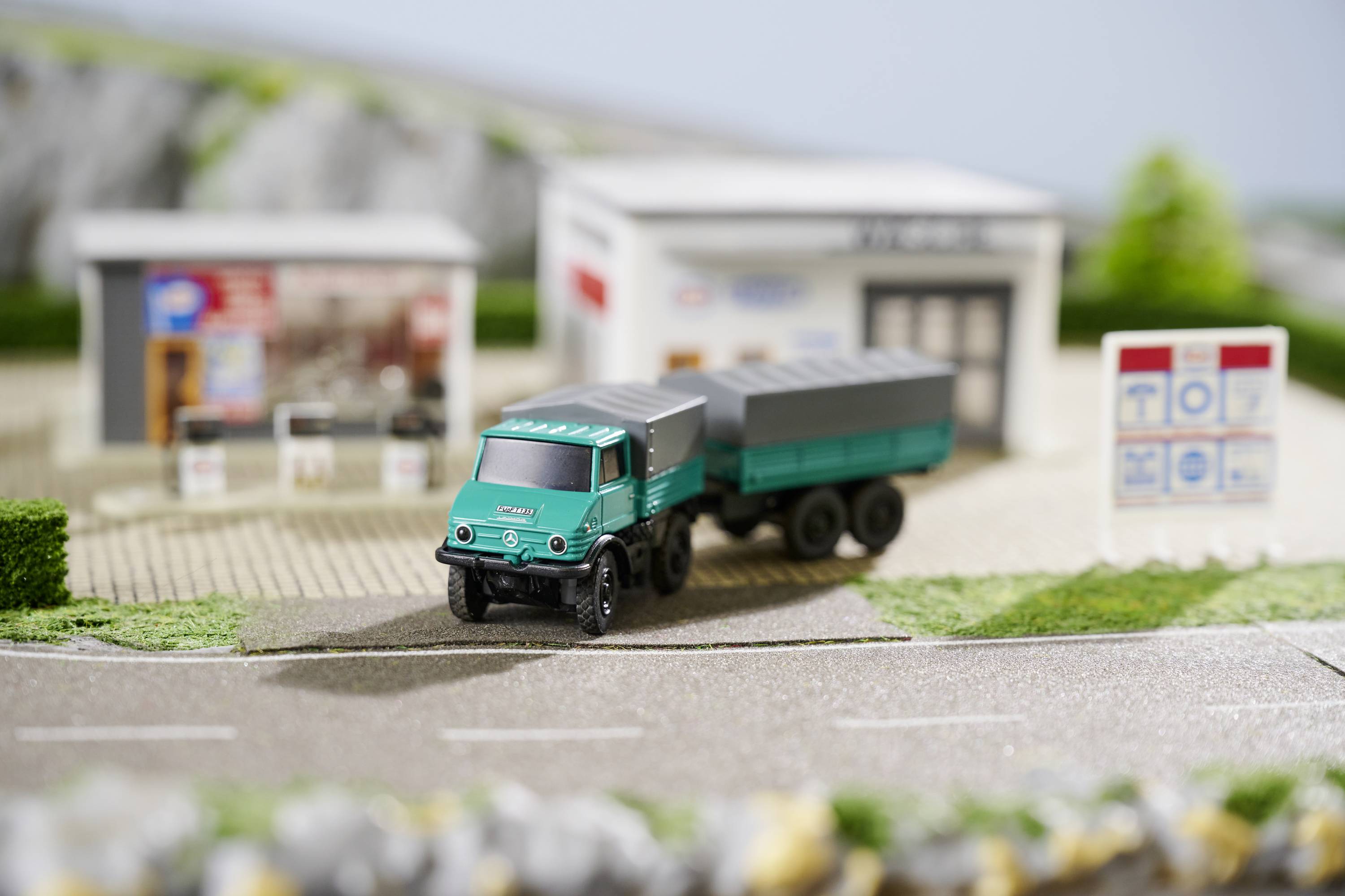 Camion jouet sur une petite route miniature devant une maison de modèles réduits avec deux bâtiments et des panneaux en arrière-plan.