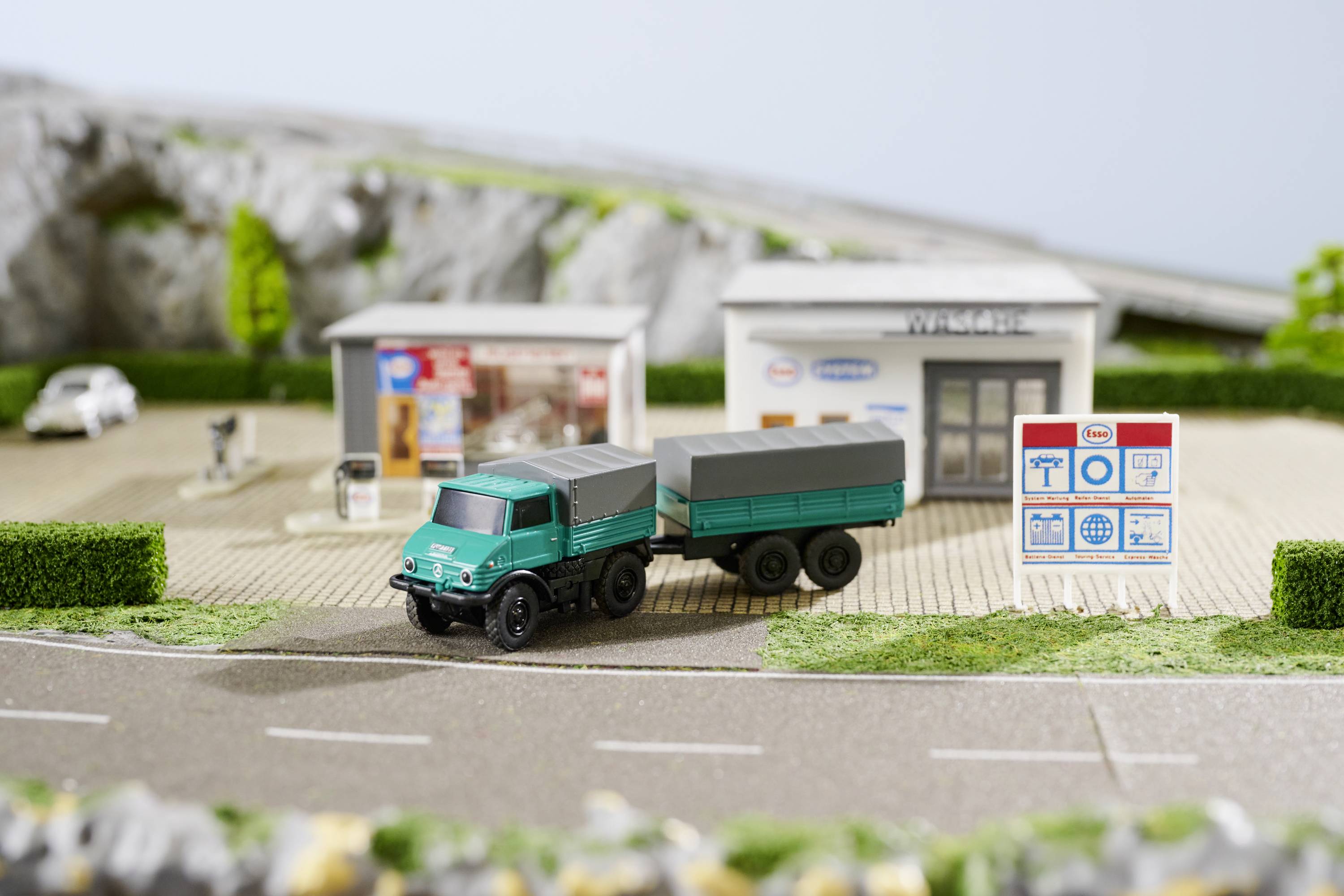 Un camion-jouet avec remorque est stationné devant une miniature d'une station-service comprenant deux bâtiments et des panneaux publicitaires en arrière-plan.
