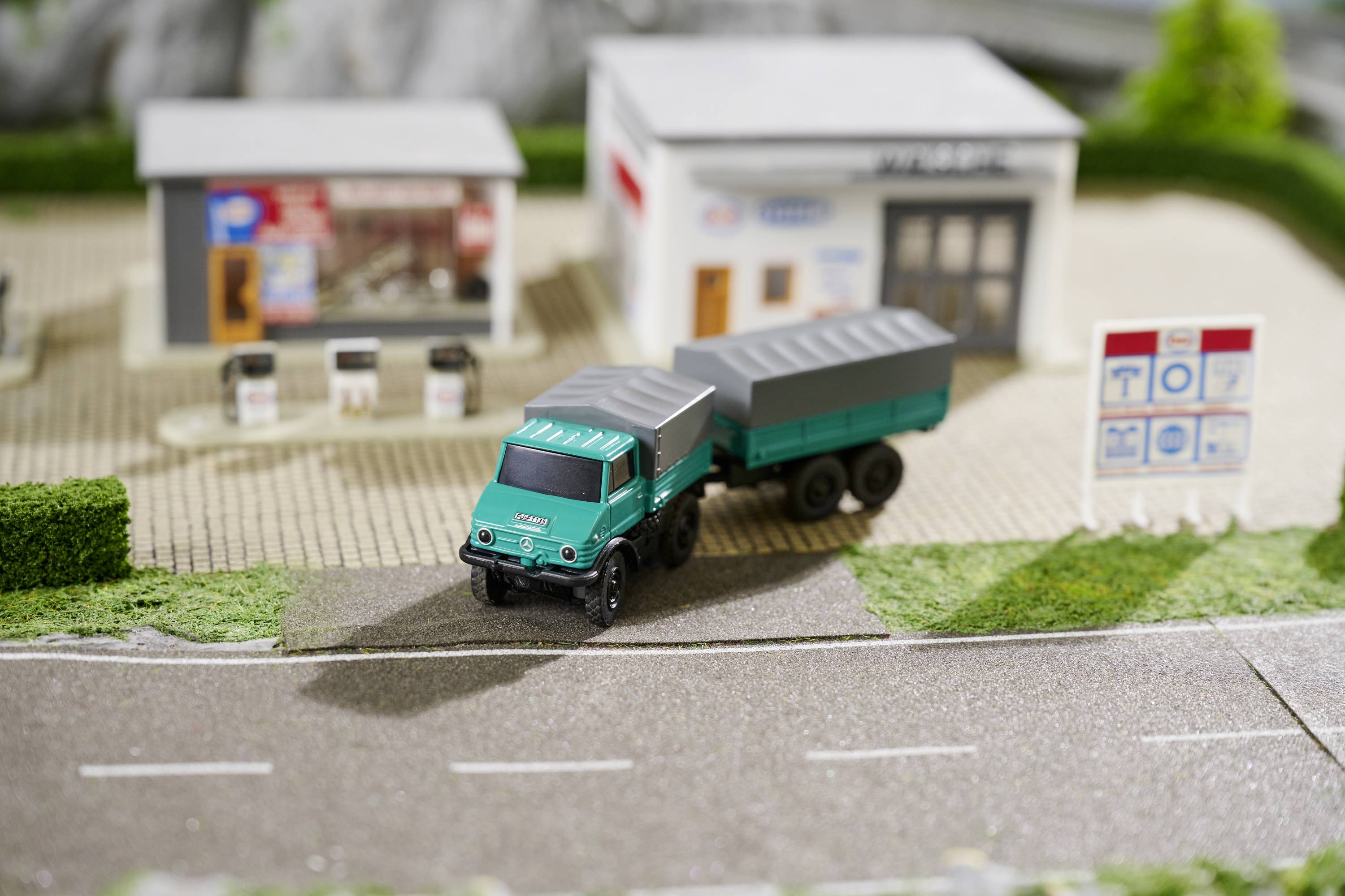 Un camion-jouet est stationné devant deux bâtiments miniatures ressemblant à des stations-service. Des panneaux de signalisation sont visibles en arrière-plan.