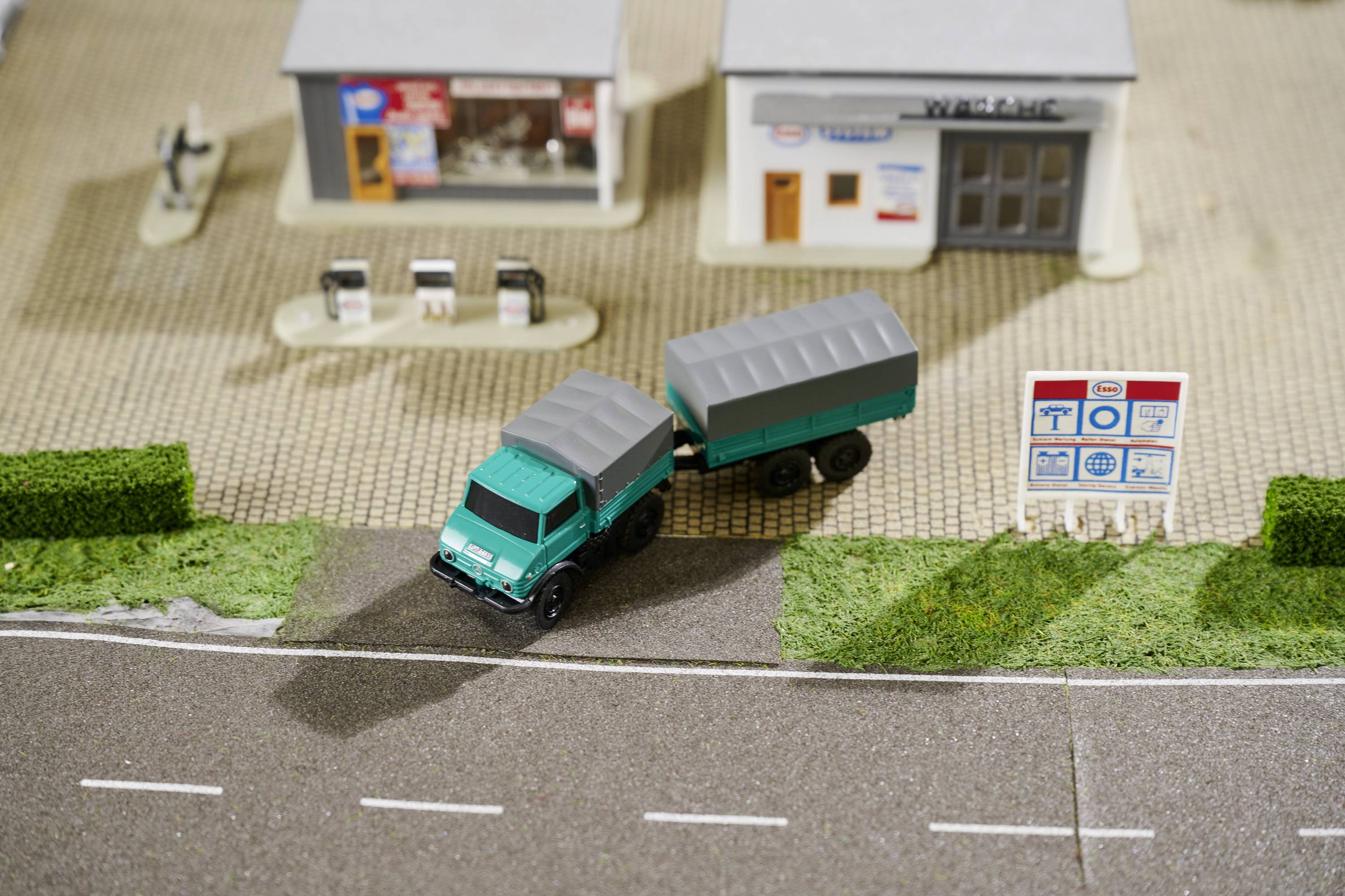 Un camion miniature quitte une route pour emprunter l'entrée d'une station-service avec deux petits bâtiments et un panneau.
