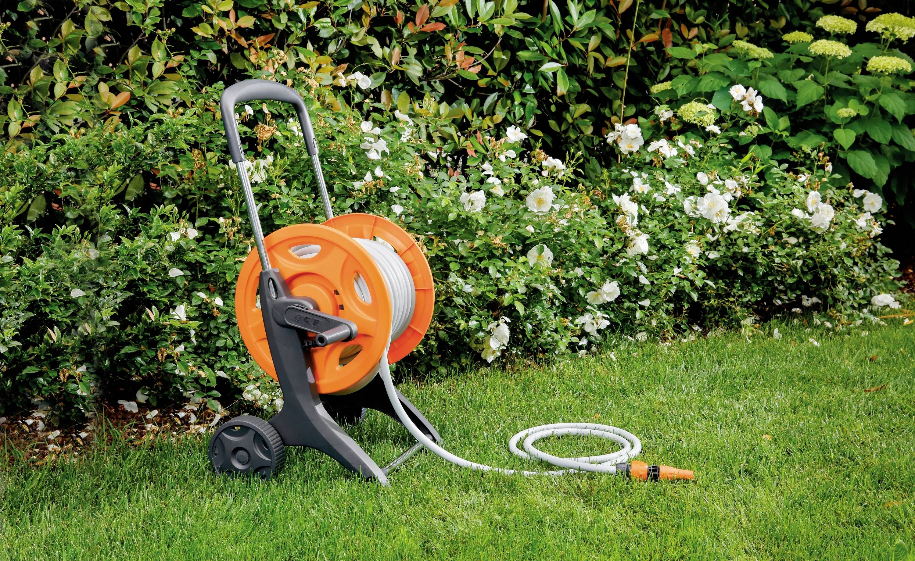 Un chariot de tuyau d'arrosage orange et gris est posé sur une pelouse verte devant une haie en fleurs, prêt à irriguer.