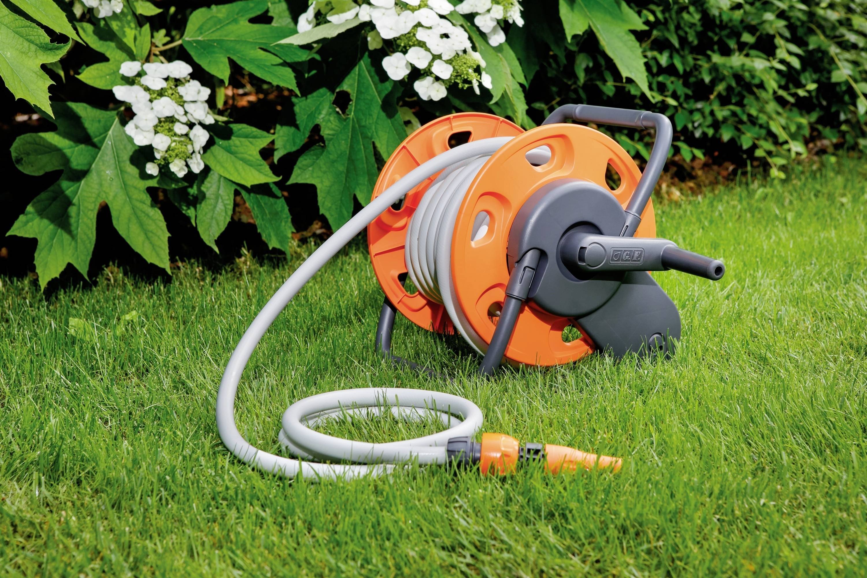 Tuyau d'arrosage enroulé sur un dévidoir orange, posé sur une pelouse verte devant des arbustes en fleurs. Scène de jardin dans un environnement estival.