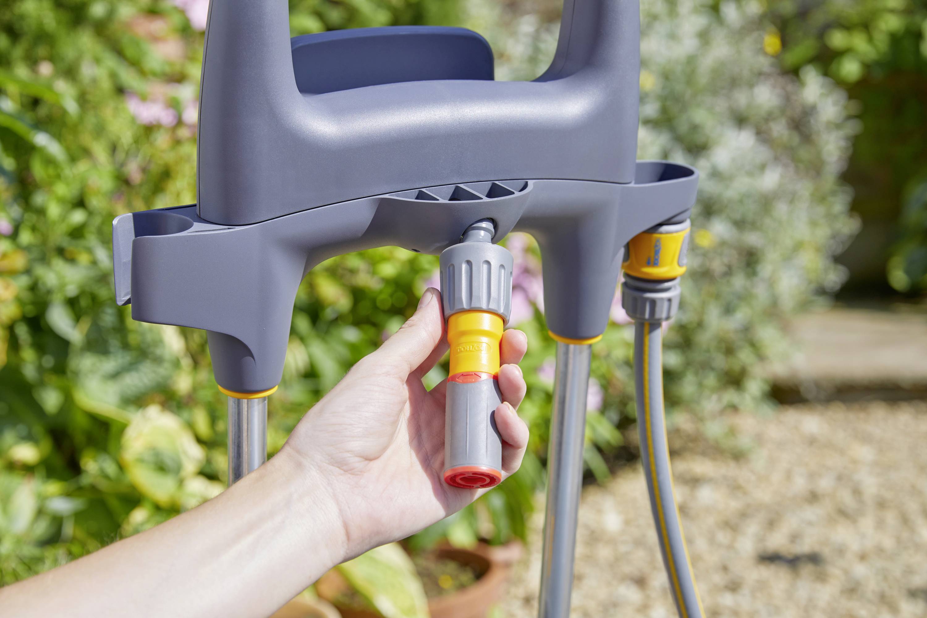 Une main tient un connecteur de tuyau relié à un tuyau d'arrosage et à un support. Des plantes sont visibles en arrière-plan.