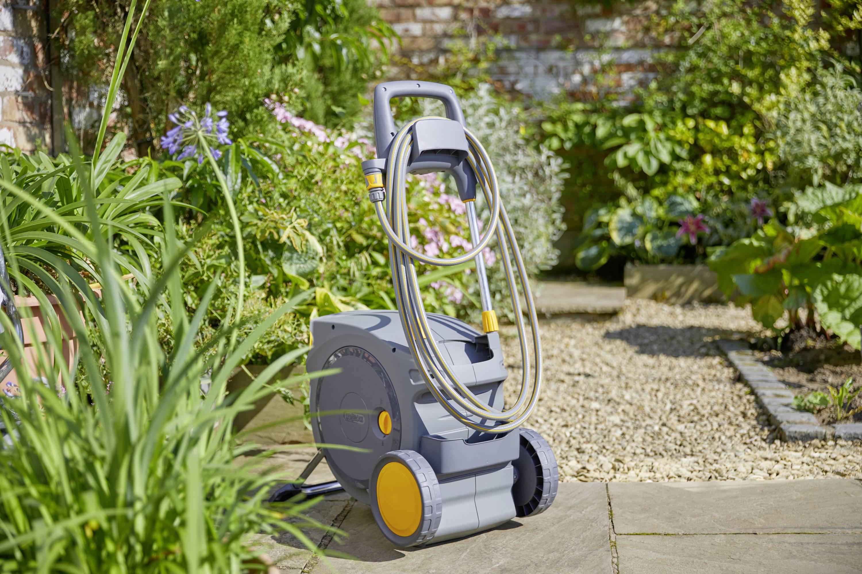 Un chariot de tuyau d'eau mobile est installé sur une terrasse de jardin, entouré de plantes vertes et de fleurs. Idéal pour l'arrosage du jardin.