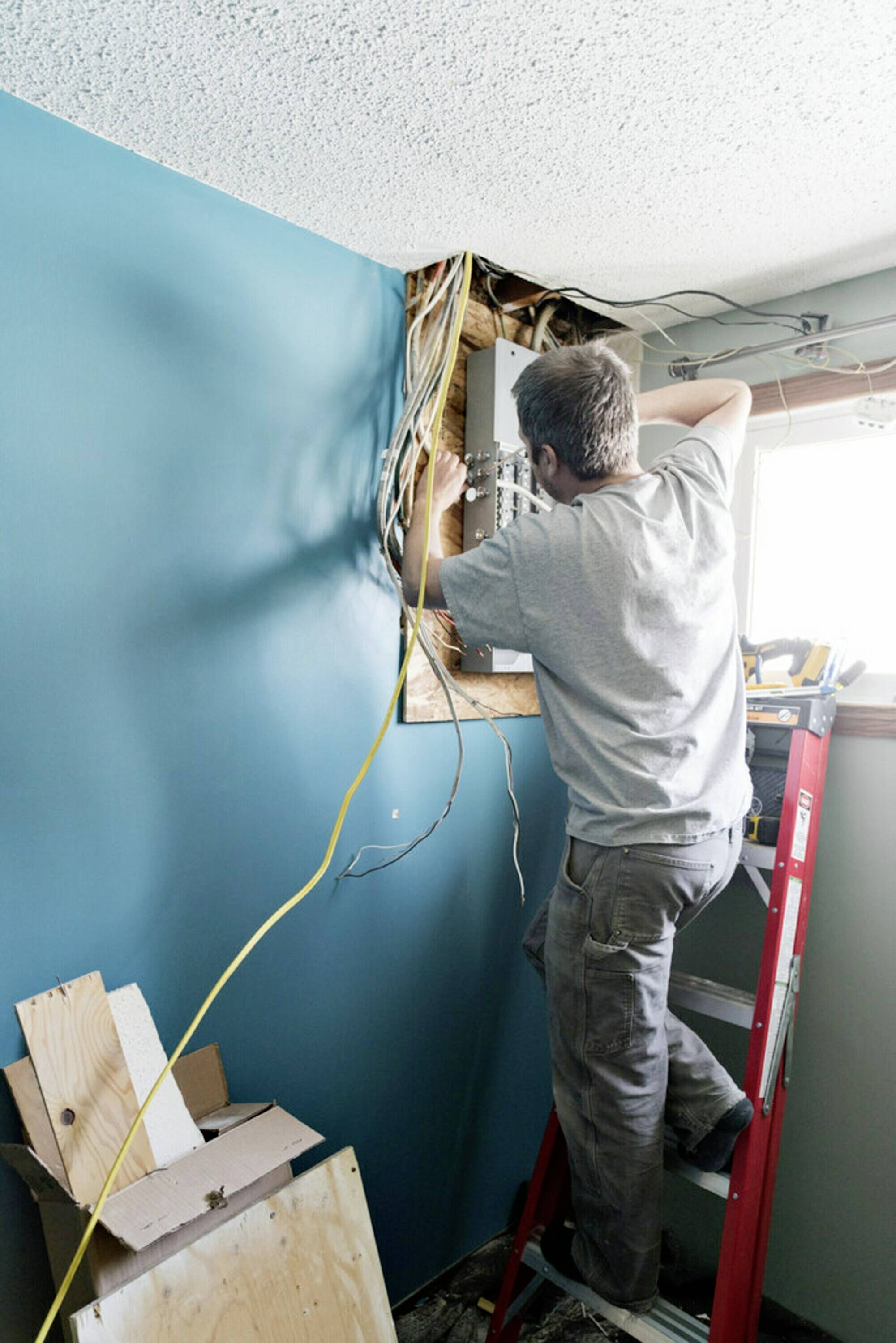 Une personne répare des câbles électriques dans un mur. Elle est debout sur une échelle et travaille avec concentration pour réparer le dommage.