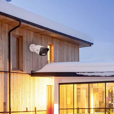 Maison en bois avec de la neige sur le toit, surveillée par une caméra de sécurité extérieure, éclairée par une lumière intérieure chaleureuse.