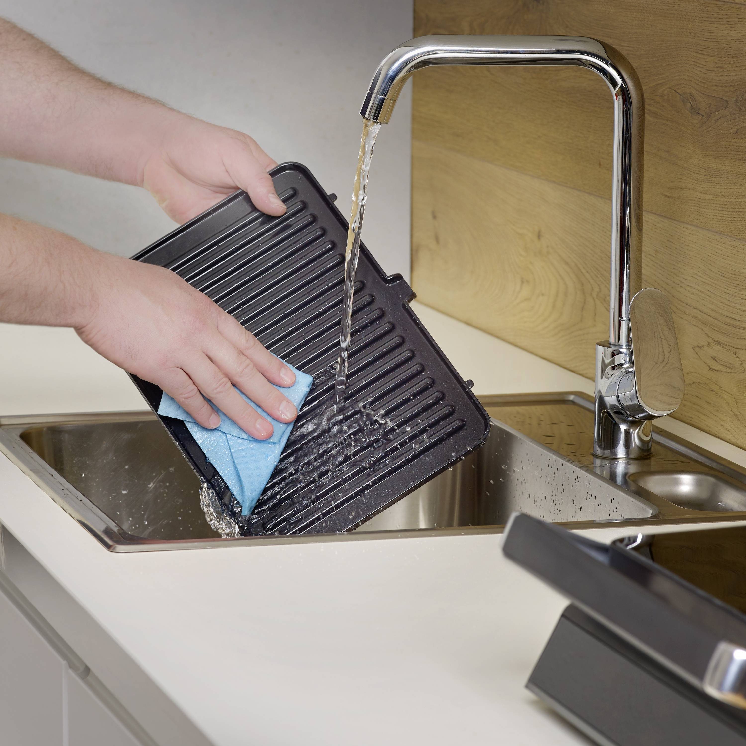 Une personne nettoie une plaque de grill avec une éponge bleue dans un évier, pendant que l'eau coule du robinet.