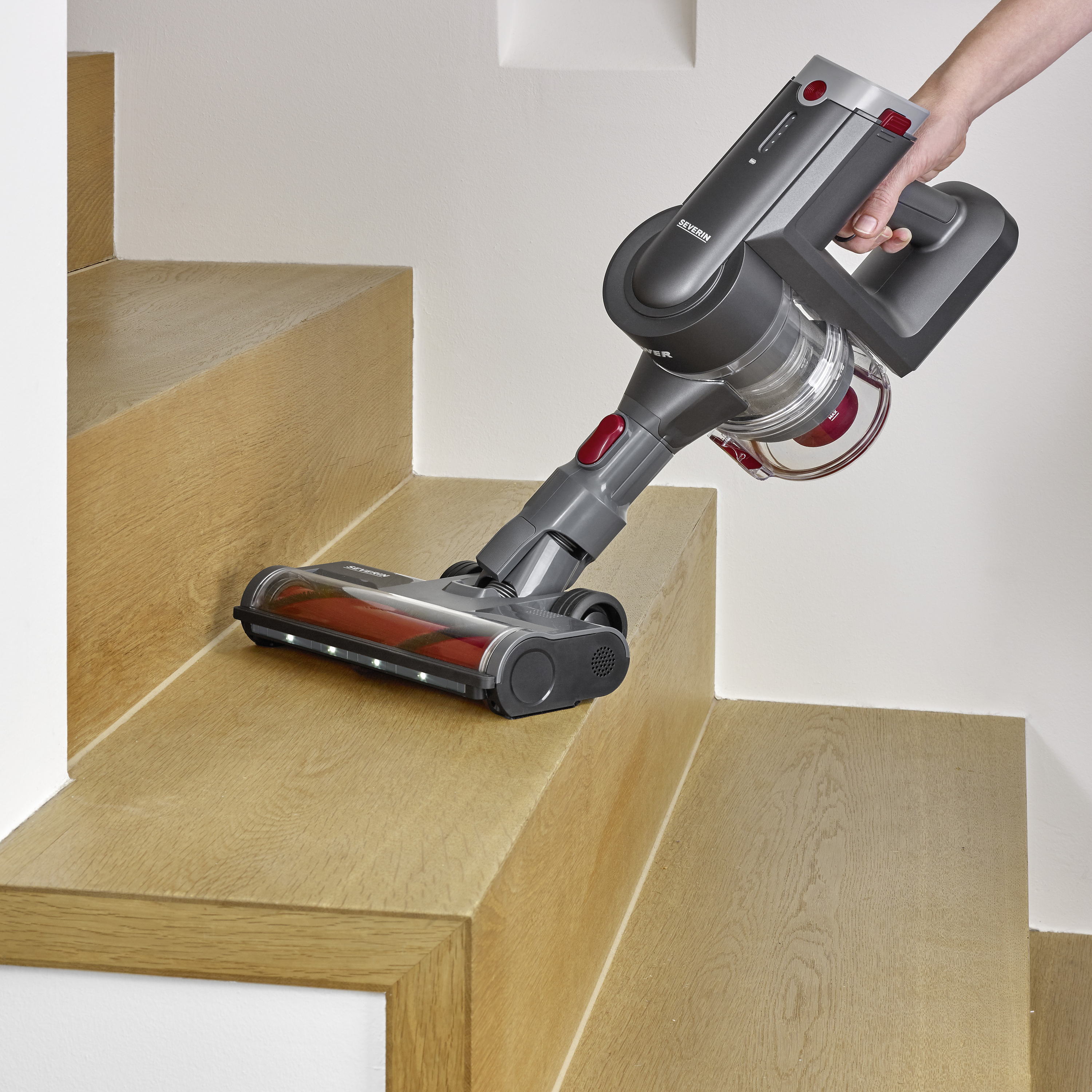 Un aspirateur sans fil nettoie un escalier en bois. Une main tient la poignée de l'aspirateur, tandis que la brosse passe sur les marches.