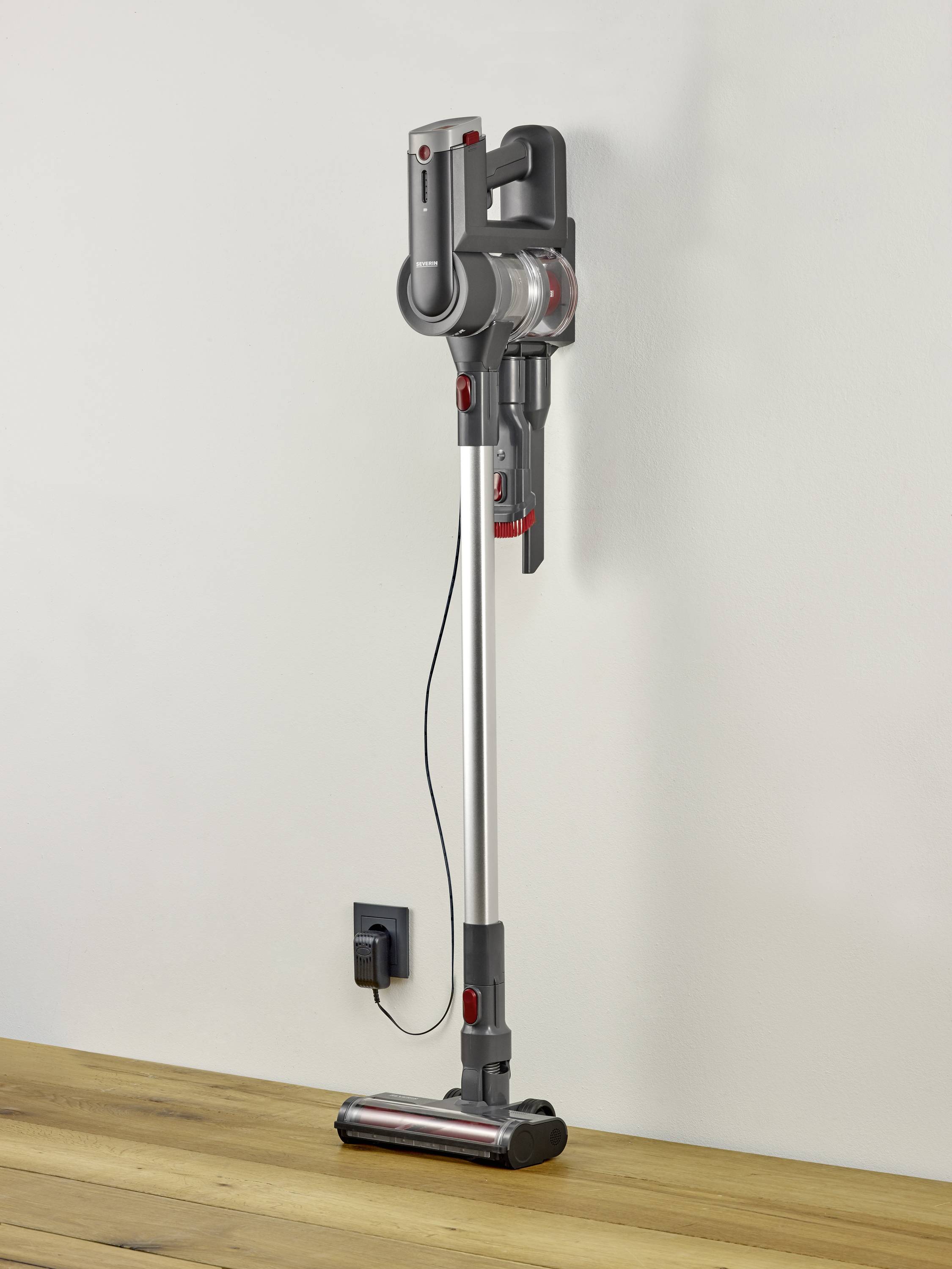 Un aspirateur sans fil est suspendu verticalement à un mur blanc. Il présente un design moderne avec des accents gris et rouges.