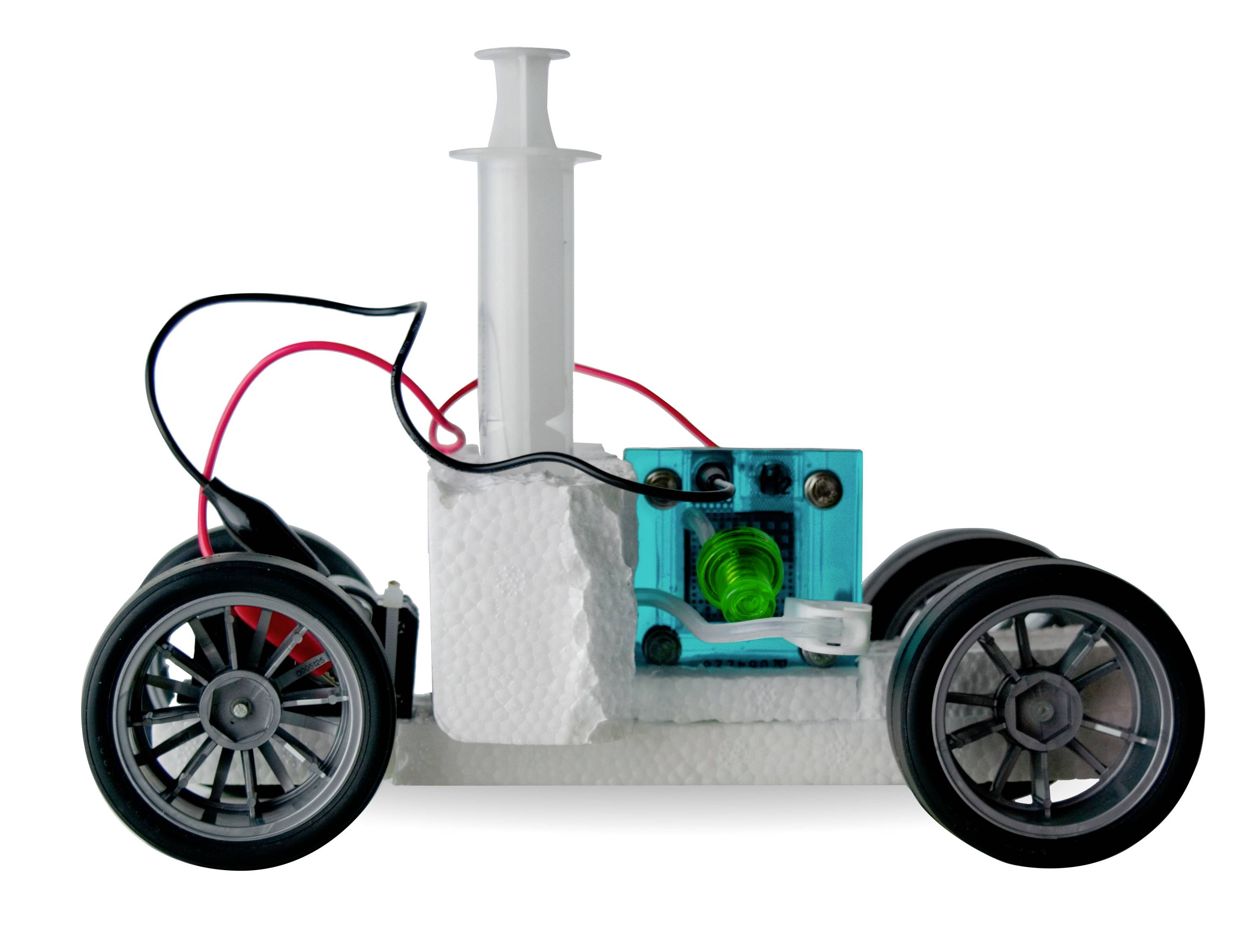Une petite voiture artisanale à quatre roues, avec un cylindre de seringue, des câbles et un petit moteur sur une base blanche.