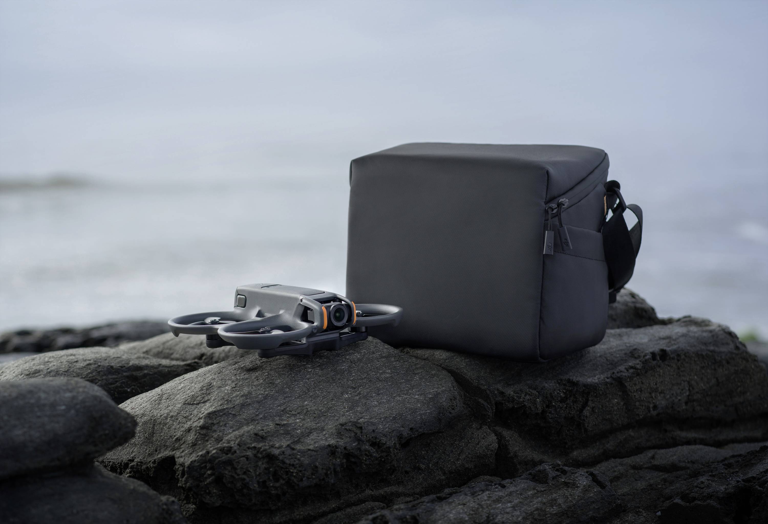 Un drone compact posé à côté d'un petit sac sombre sur des rochers dans un lieu côtier brumeux.