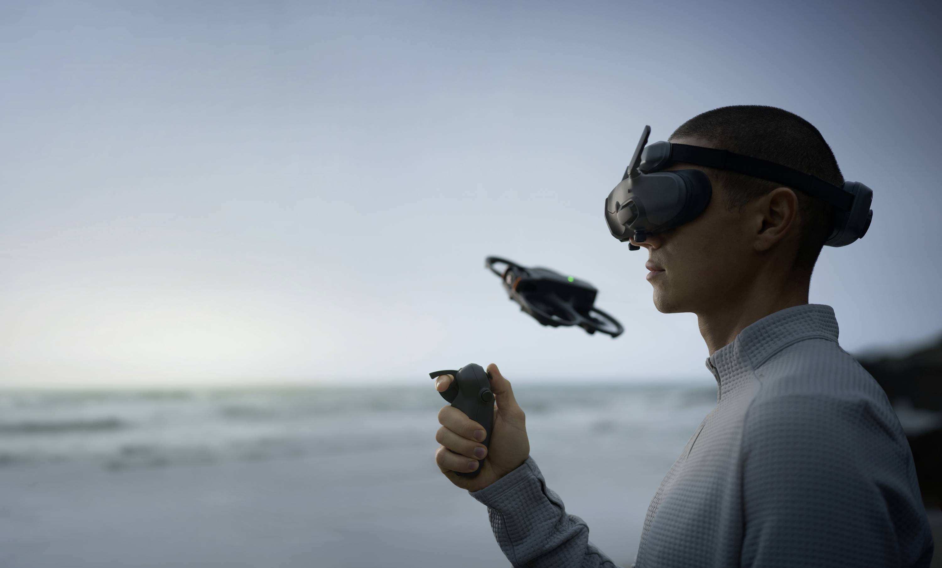 Une personne portant un casque de réalité virtuelle pilote un drone sur une plage à l'aide d'une manette de contrôle. L'horizon océanique s'étend en arrière-plan.