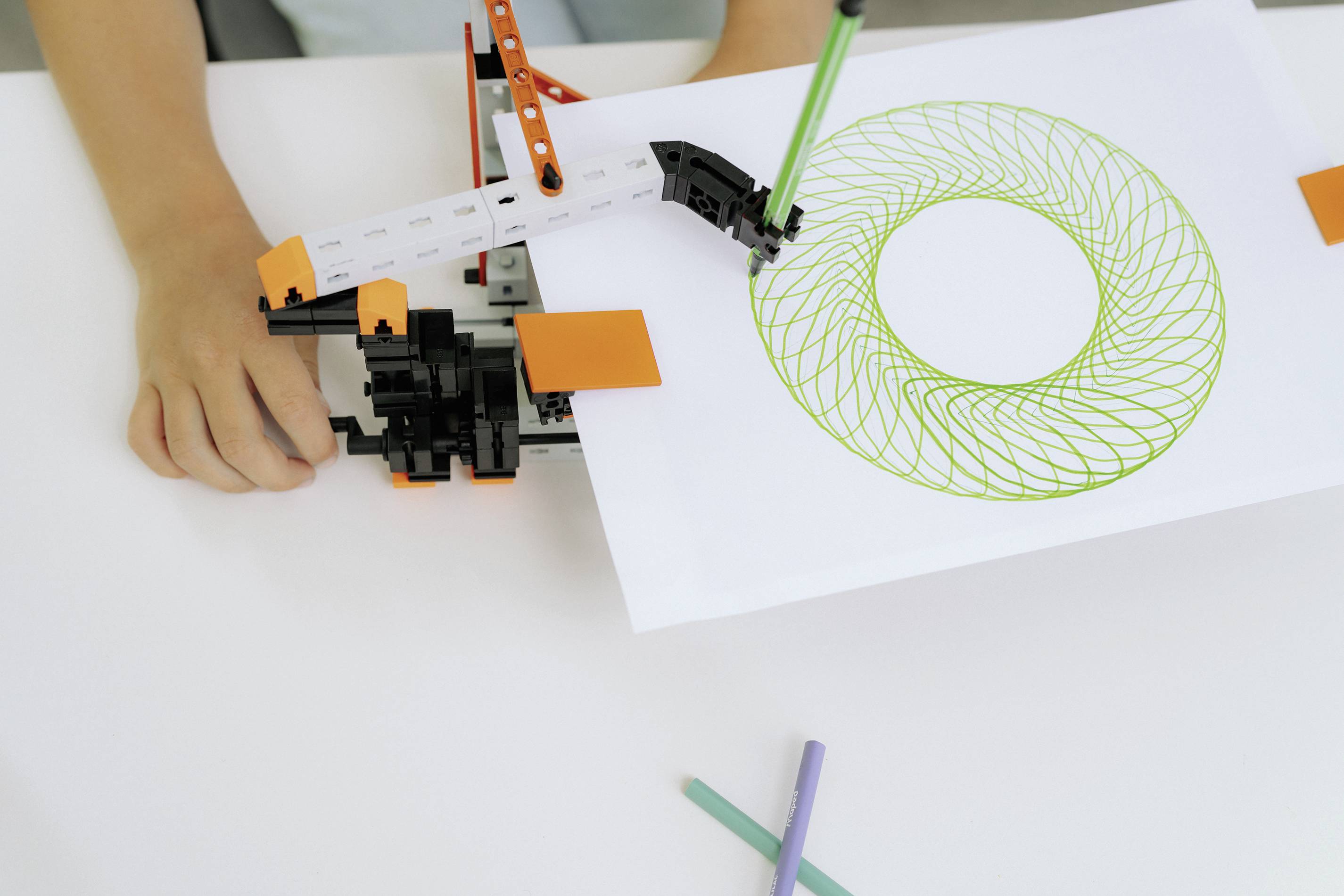 Un enfant dessine un motif circulaire vert sur du papier à l'aide d'un appareil mécanique. Quelques crayons colorés sont posés à côté.