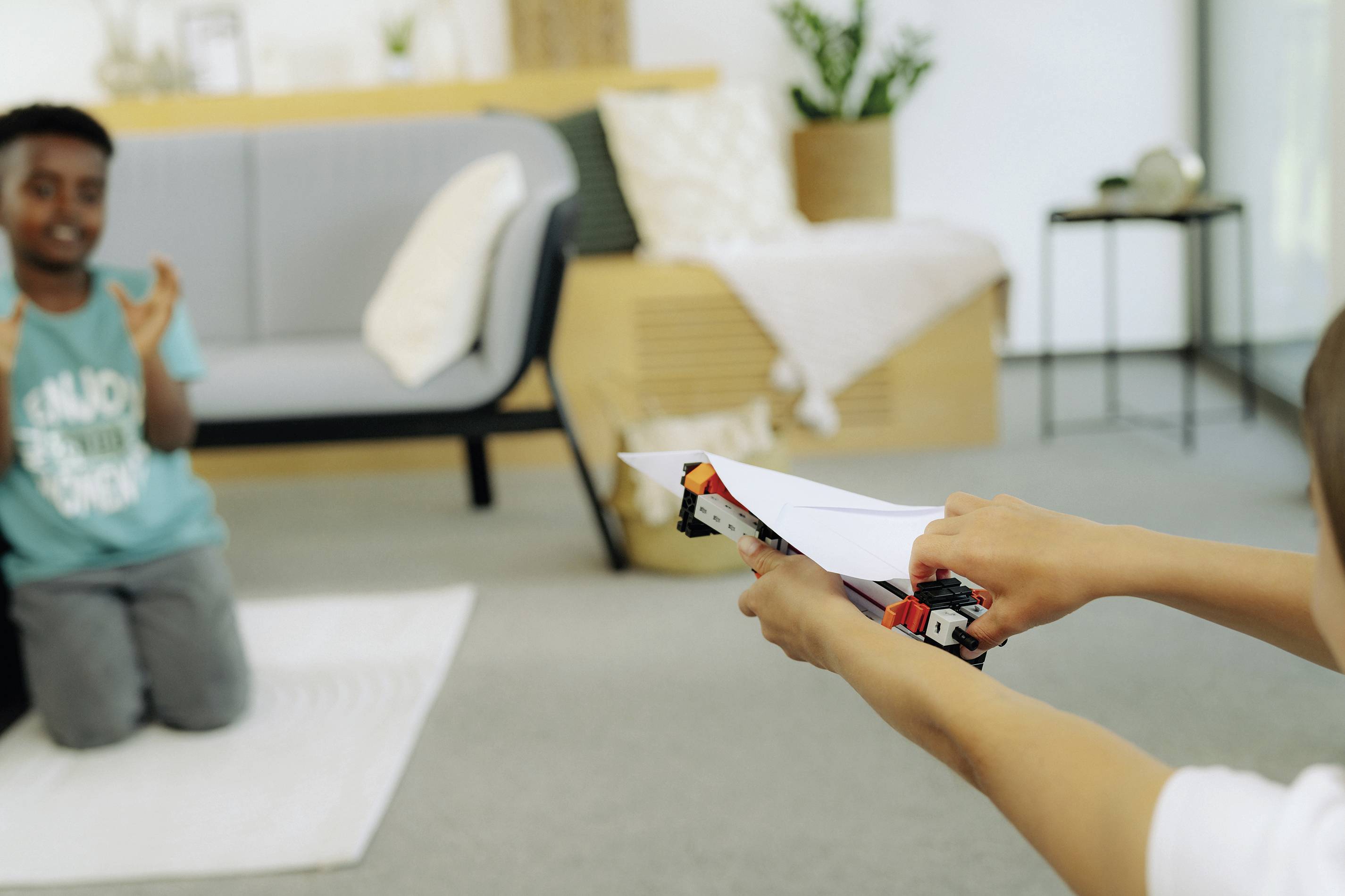 Deux enfants jouent avec des avions en papier faits maison dans le salon. Un enfant est à genoux sur le sol, l'autre lance un avion.