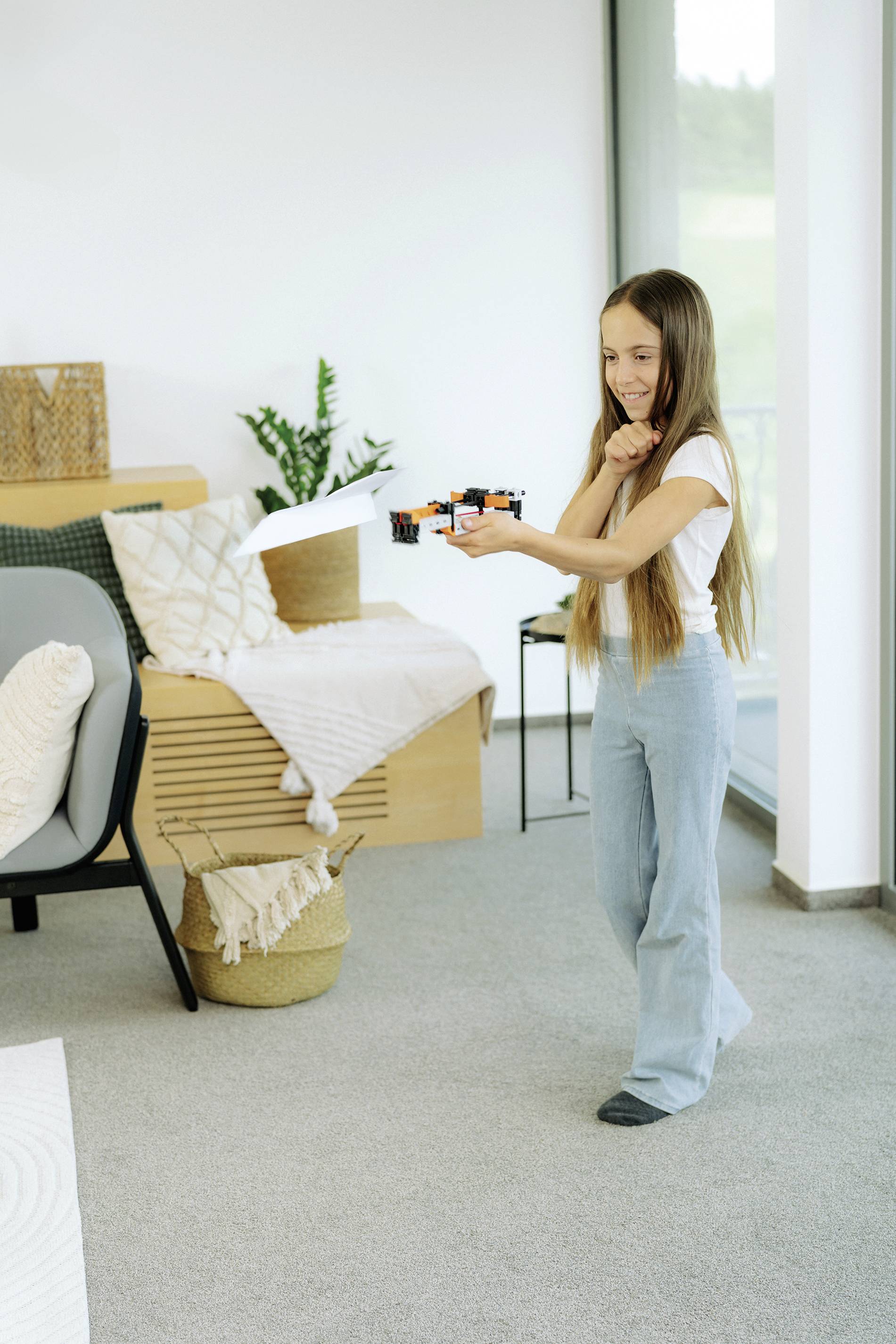 Un enfant dans un salon contrôle un petit drone à l'aide d'une télécommande. Des meubles et des plantes sont visibles en arrière-plan.