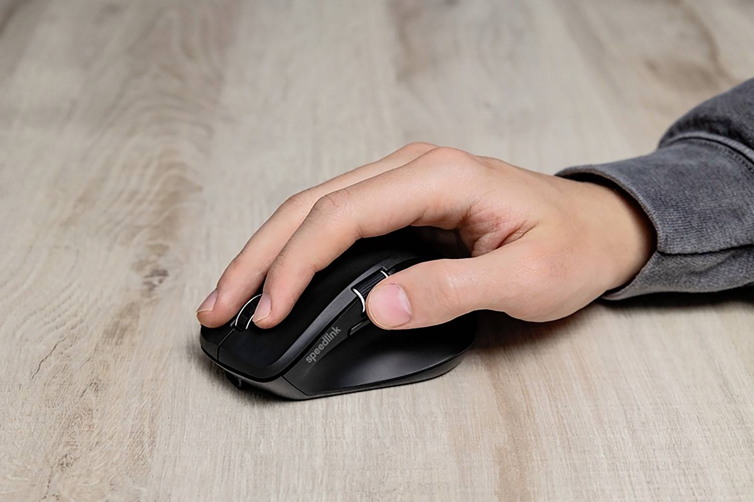 Une main droite tient une souris d'ordinateur noire sur une table en bois.