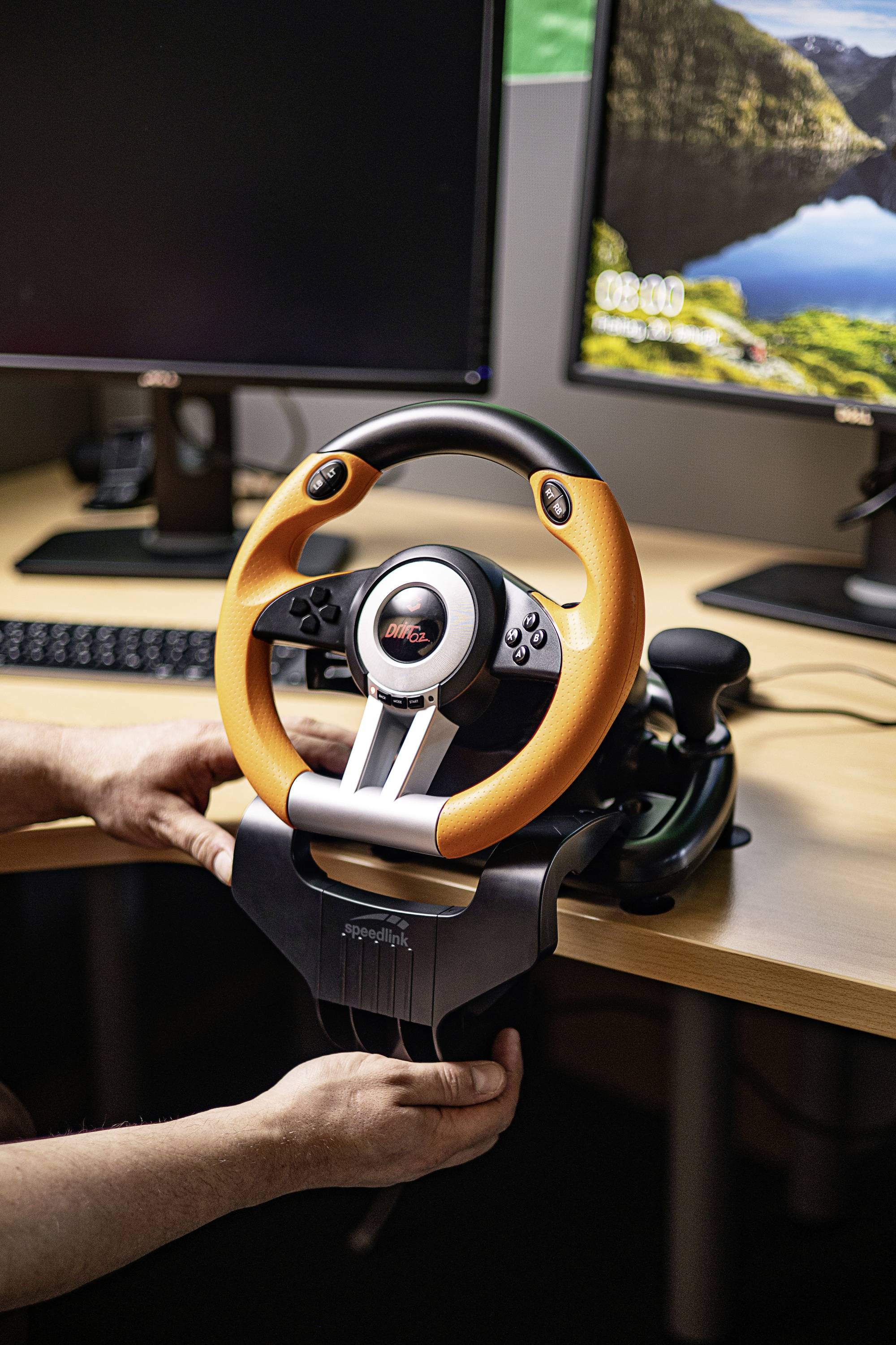 Une personne installe un volant de jeu orange sur un bureau devant deux écrans.