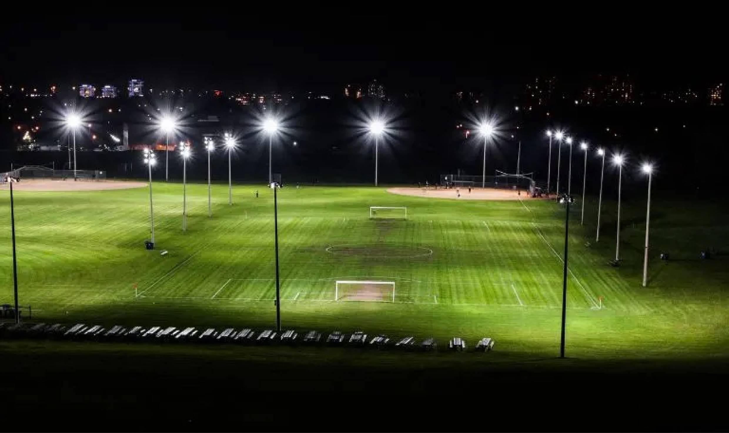 Un terrain de football éclairé la nuit, avec des buts, entouré de pylônes de projecteurs. Des lumières urbaines sont visibles en arrière-plan.