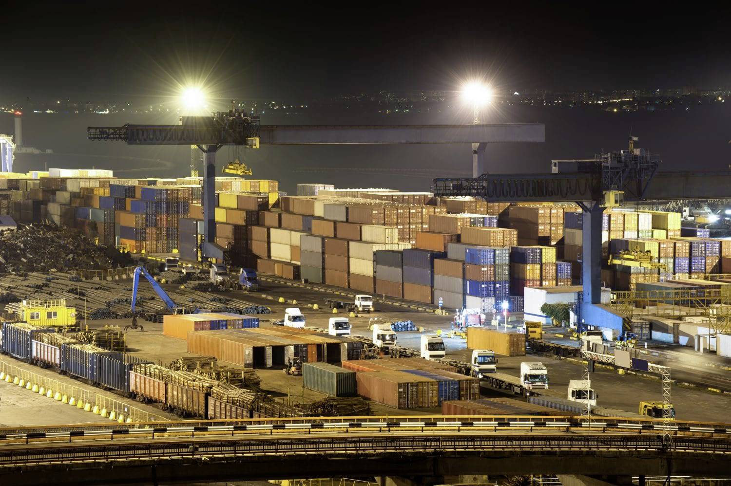 Un port nocturne avec des grues illuminées et des conteneurs de fret empilés. Plusieurs camions se déplacent entre les conteneurs.