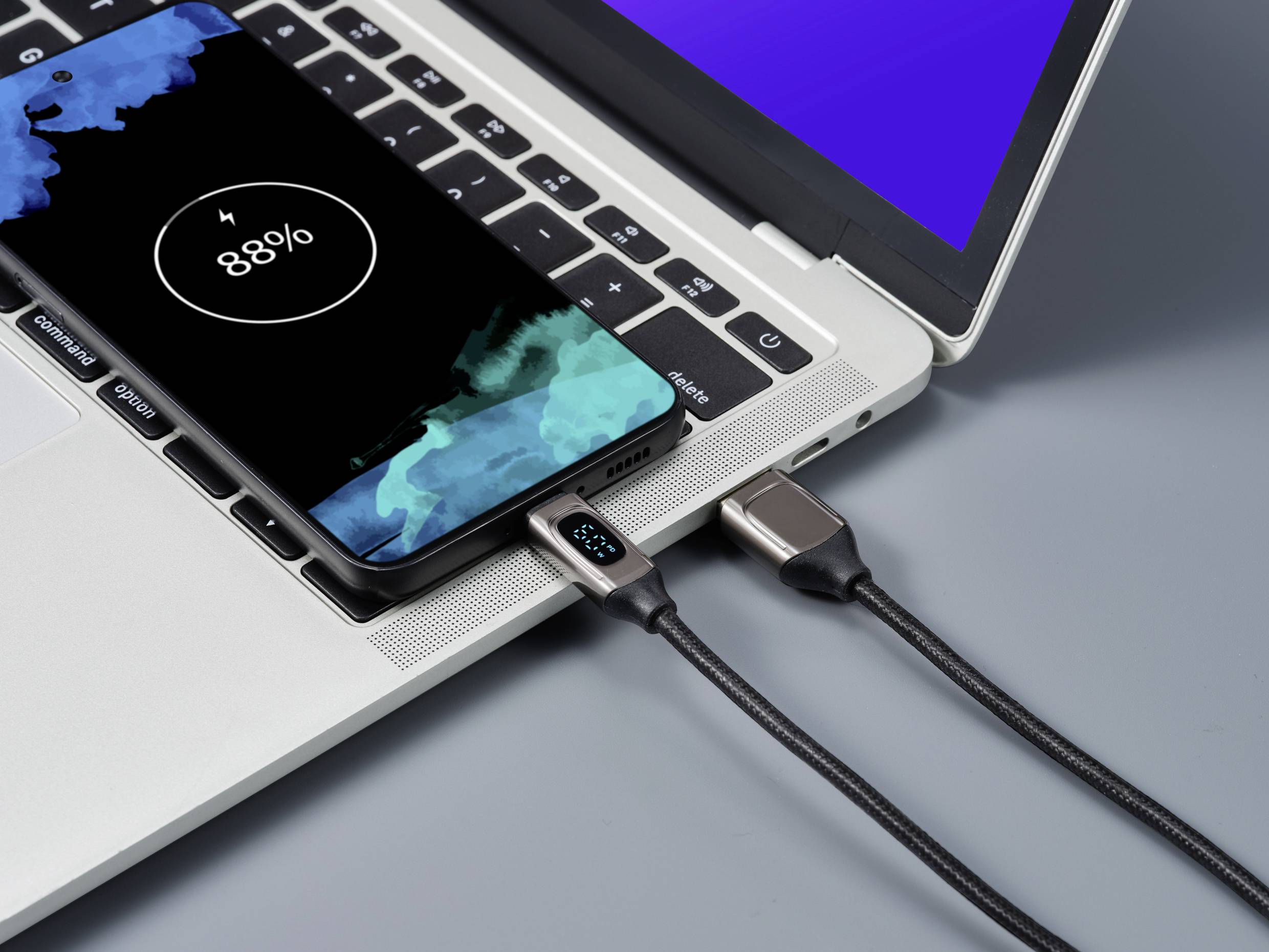 Un smartphone se charge sur un ordinateur portable. L'écran affiche '88%'. Un câble de charge relie le téléphone à l'ordinateur portable, dont l'écran est bleu.