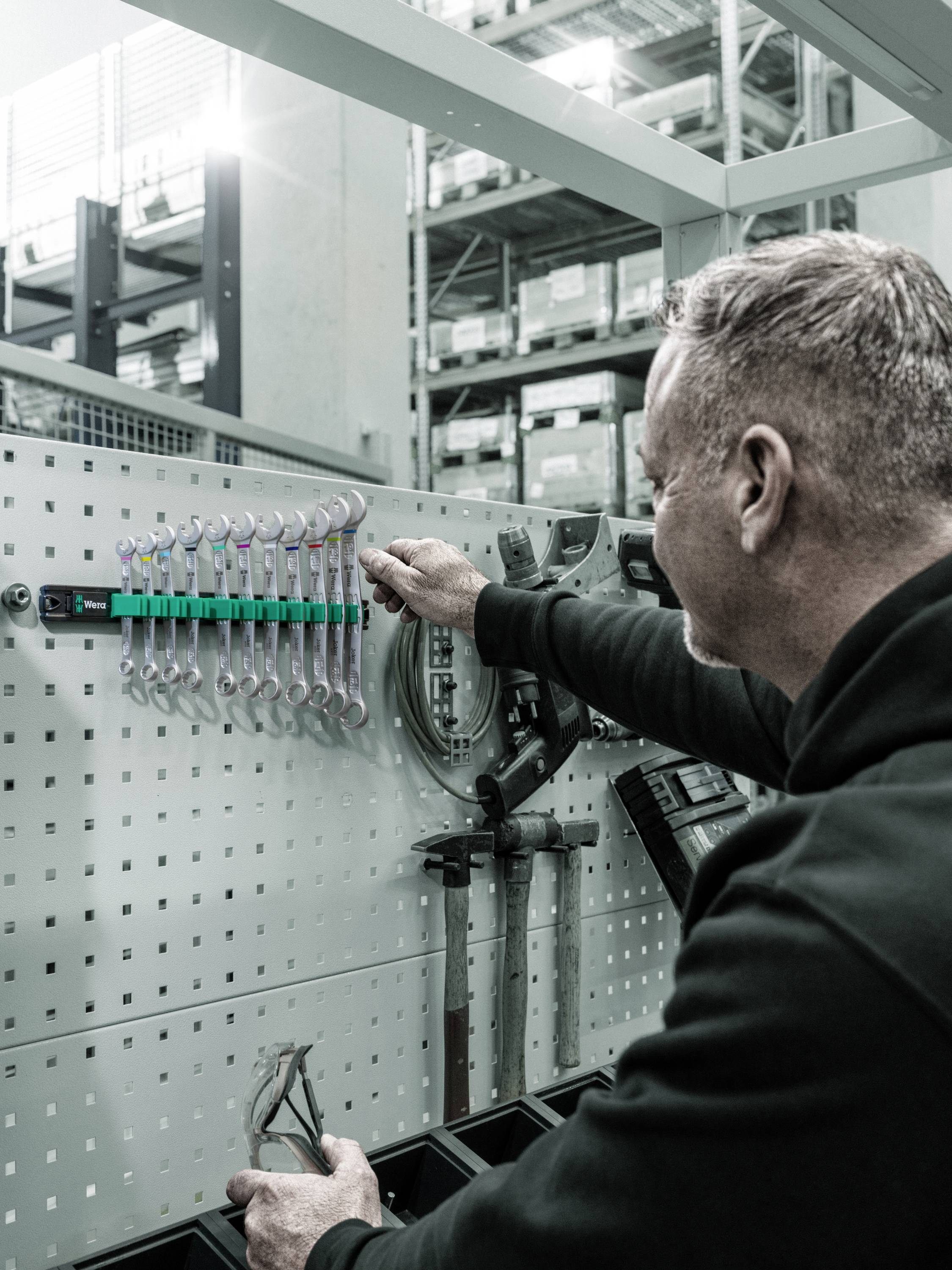 Un homme dans un entrepôt tend la main vers une clé à molette sur une barre à outils, avec des étagères remplies de caisses en arrière-plan.