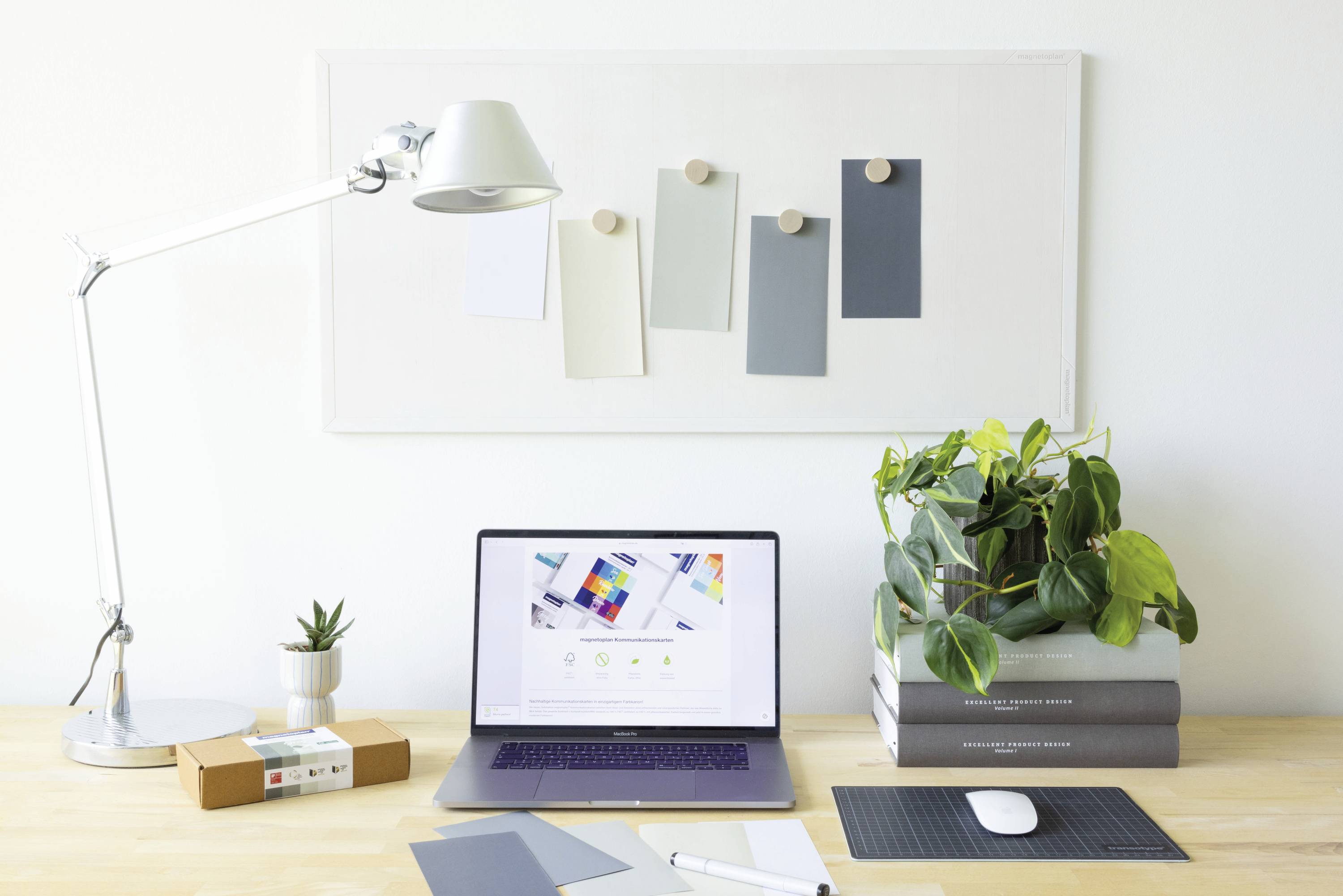Un bureau bien rangé avec un ordinateur portable, des plantes, une pile de livres, une lampe de bureau et des cartes de couleurs au mur pour un environnement de travail créatif.