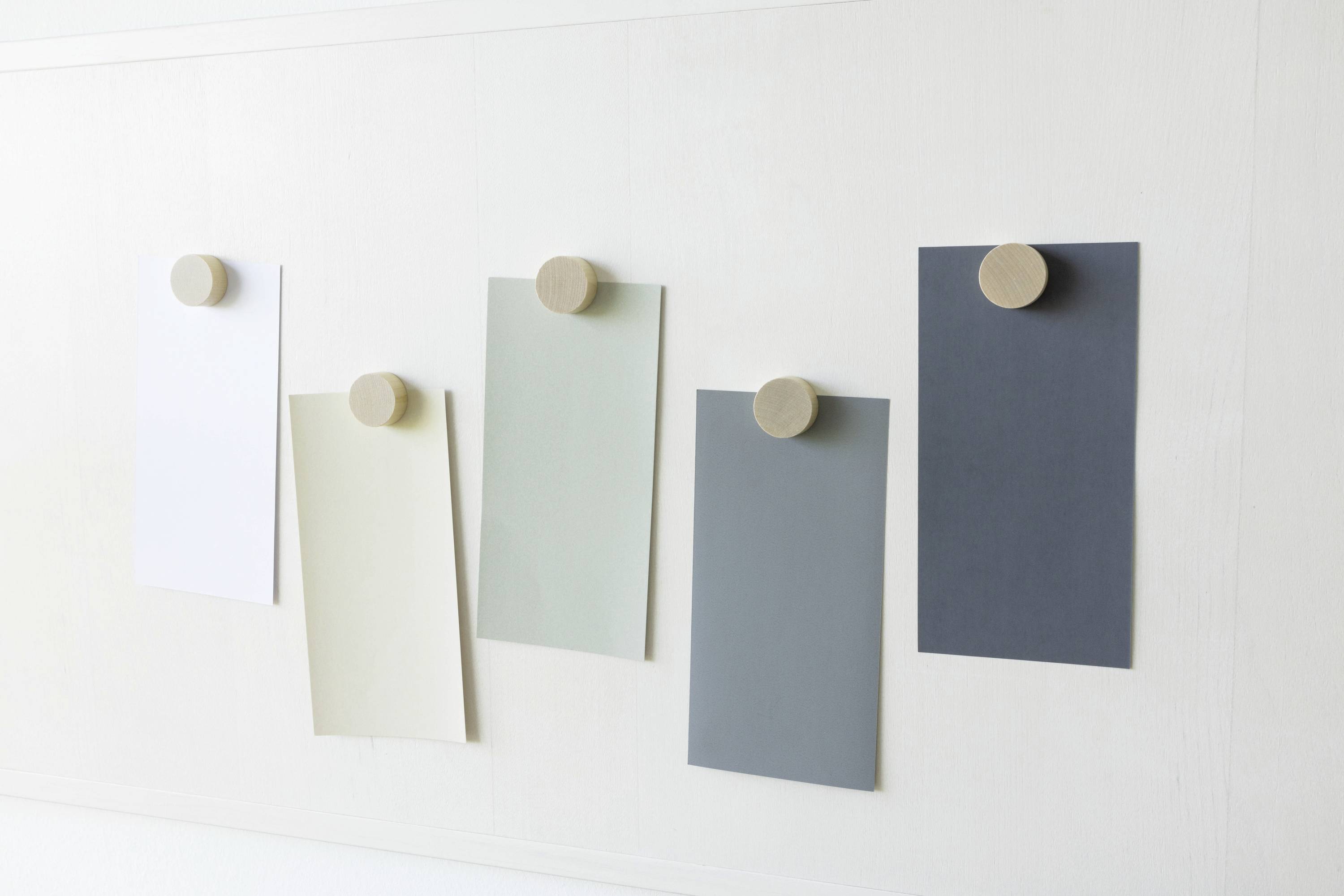 Cinq feuilles de papier colorées dans différentes nuances de gris sont fixées à un mur blanc à l'aide de supports magnétiques en bois ronds.