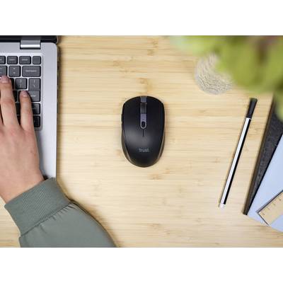 Un ordinateur portable avec une main sur le clavier, à côté une souris sans fil noire sur une table en bois, avec un stylo et un carnet de notes à proximité.