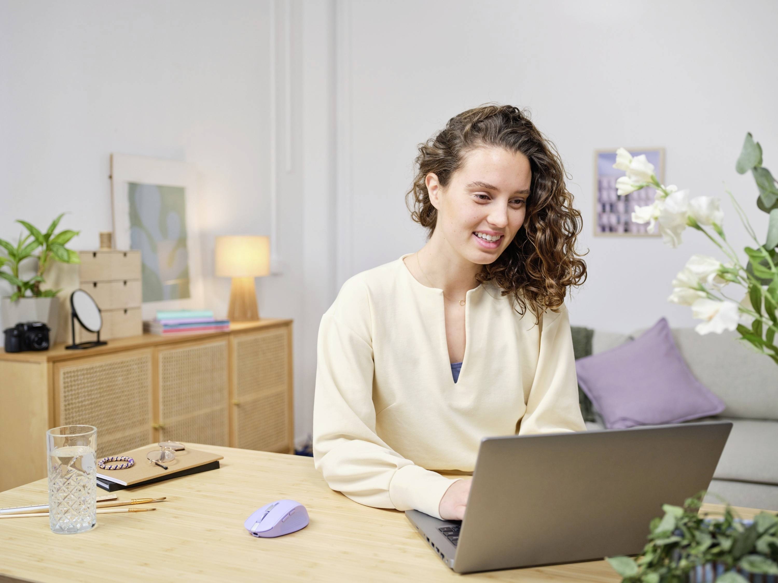 Une femme aux cheveux bouclés est assise à une table en bois et travaille sur un ordinateur portable. Des plantes et des meubles sont visibles en arrière-plan.