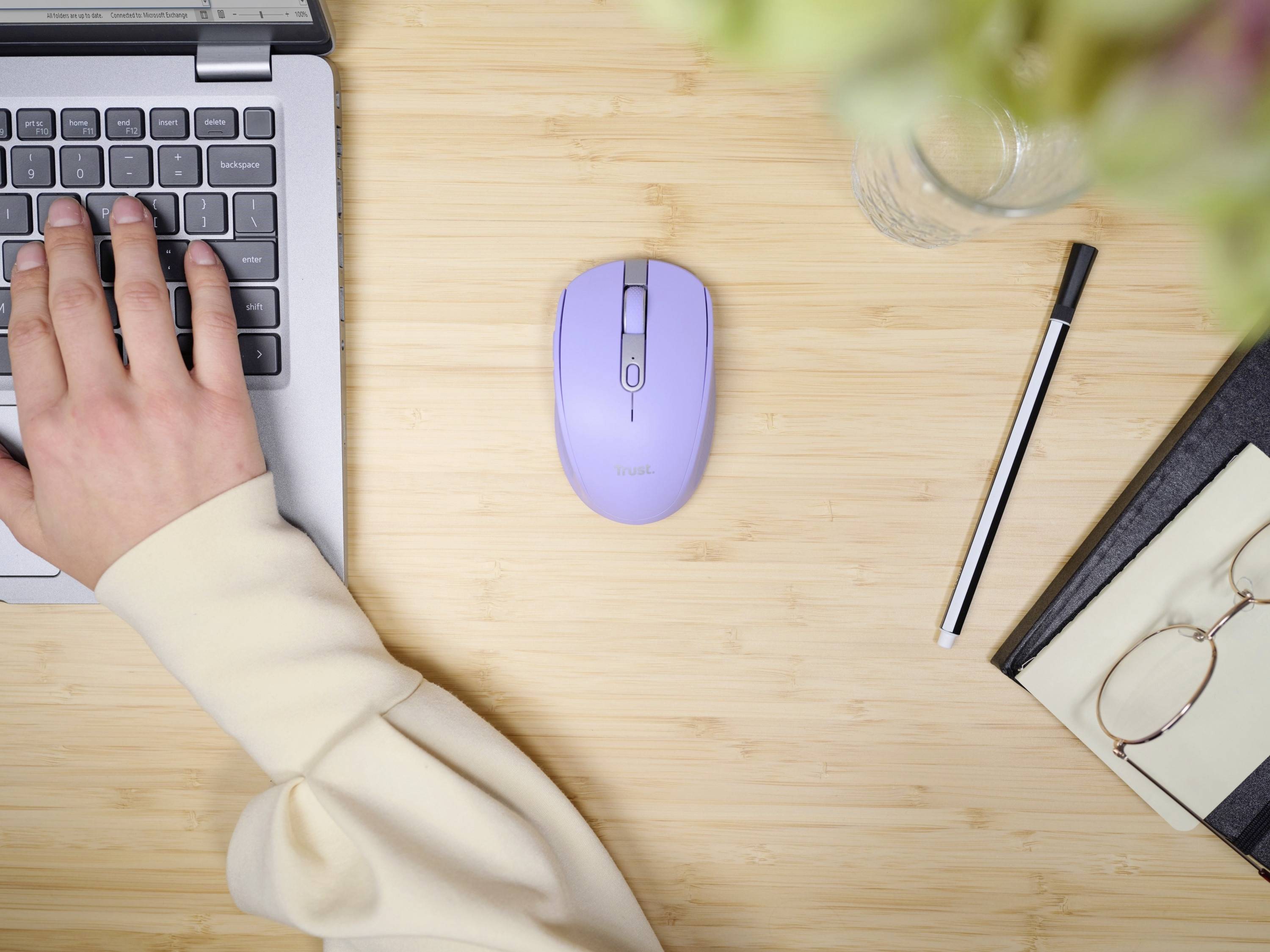 Une main tape sur le clavier d'un ordinateur portable. À côté, une souris violette, un stylo, des lunettes et un carnet de notes sont posés sur une table en bois.
