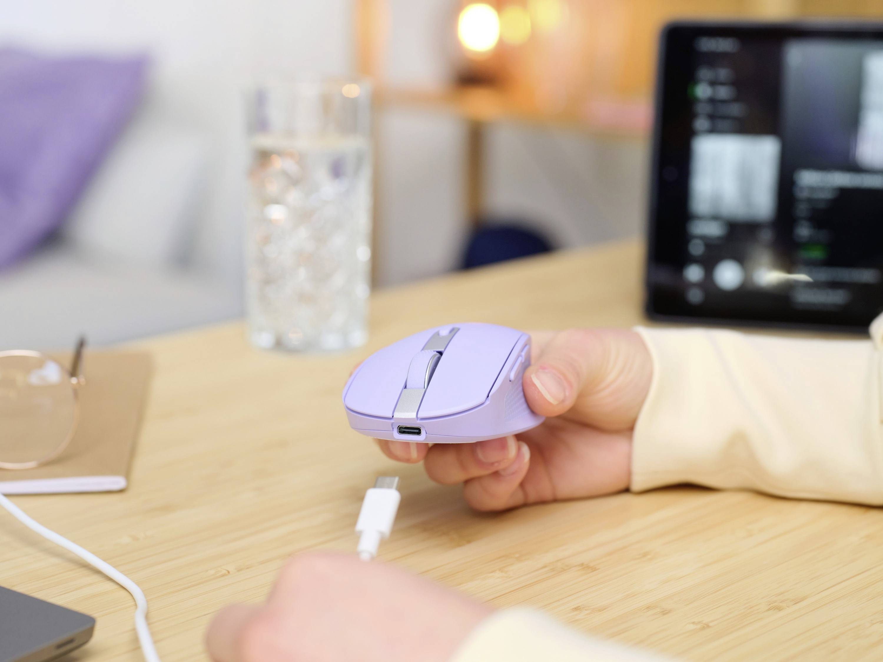 Une main tient une souris d'ordinateur violette au-dessus d'une table. Un câble USB est en train d'être branché. Un ordinateur portable et un verre d'eau sont visibles en arrière-plan.