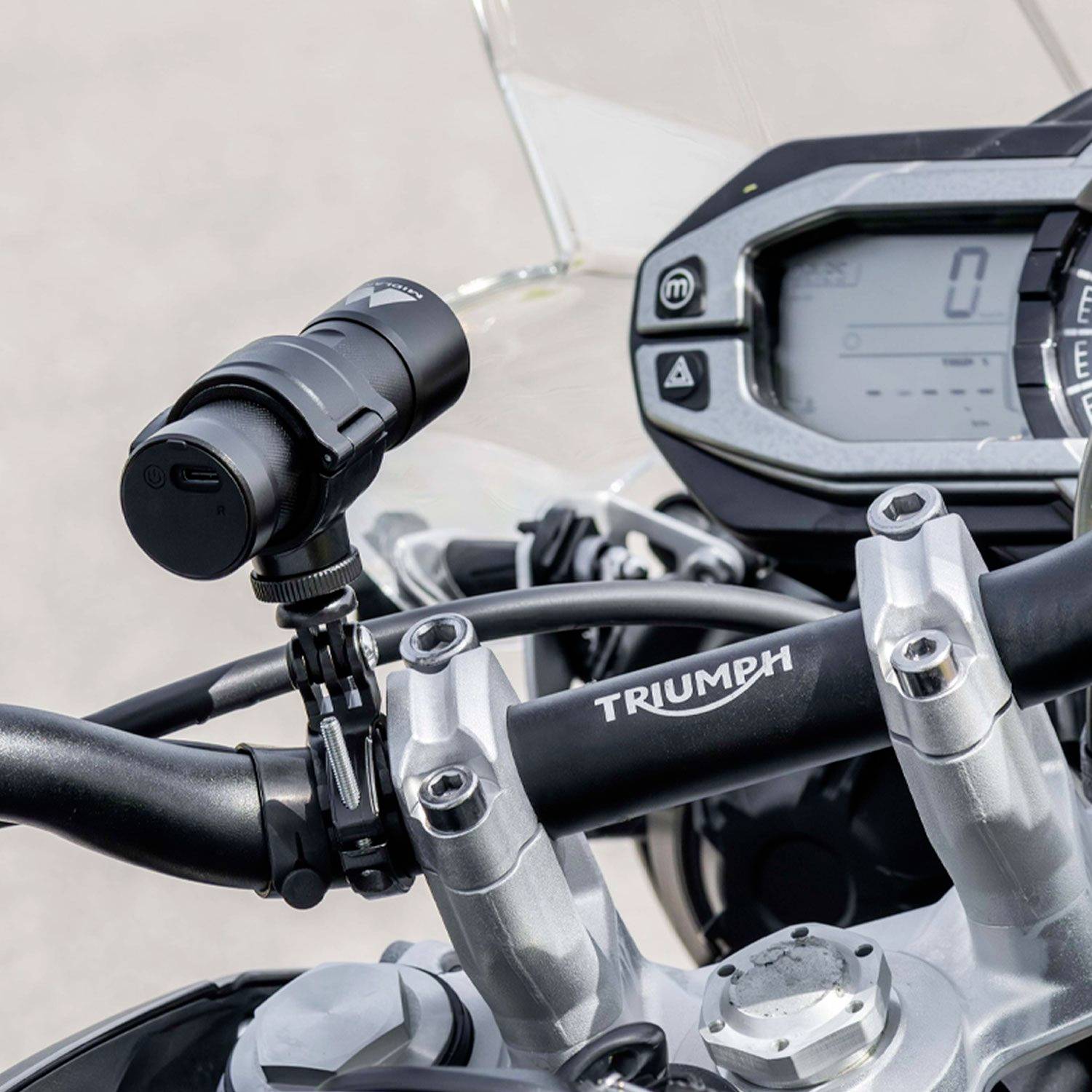 Gros plan sur le guidon d'une moto avec un dispositif technique et un compteur de vitesse numérique. Le logo Triumph est visible sur le tube de direction.