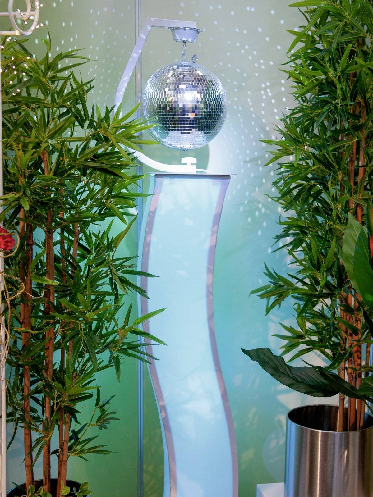 Une boule à facettes argentée est suspendue au-dessus d'une colonne éclairée, entourée de plantes vertes qui créent une atmosphère de fête ou de célébration.