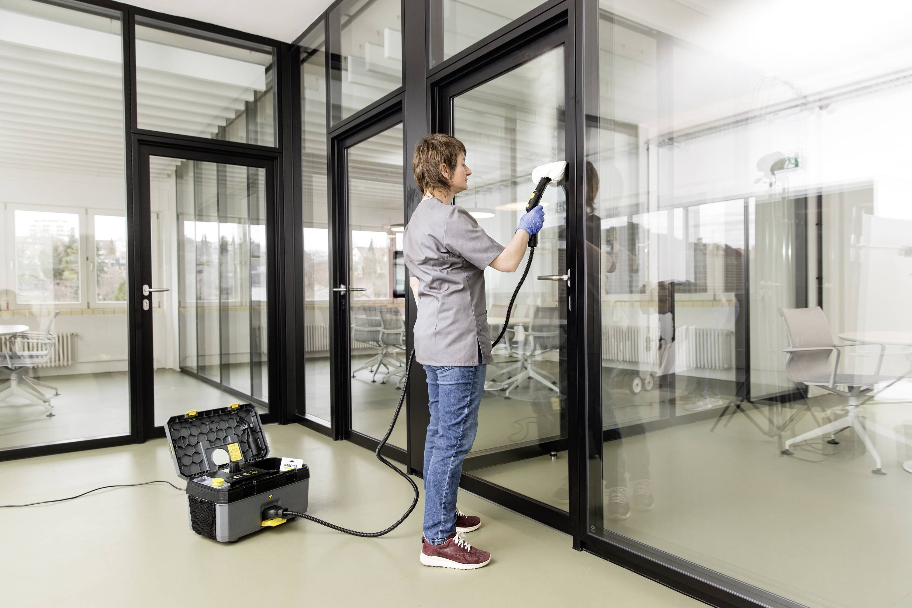 Une personne nettoie une grande paroi vitrée avec un nettoyeur vapeur dans un bureau moderne, avec des fenêtres en arrière-plan.