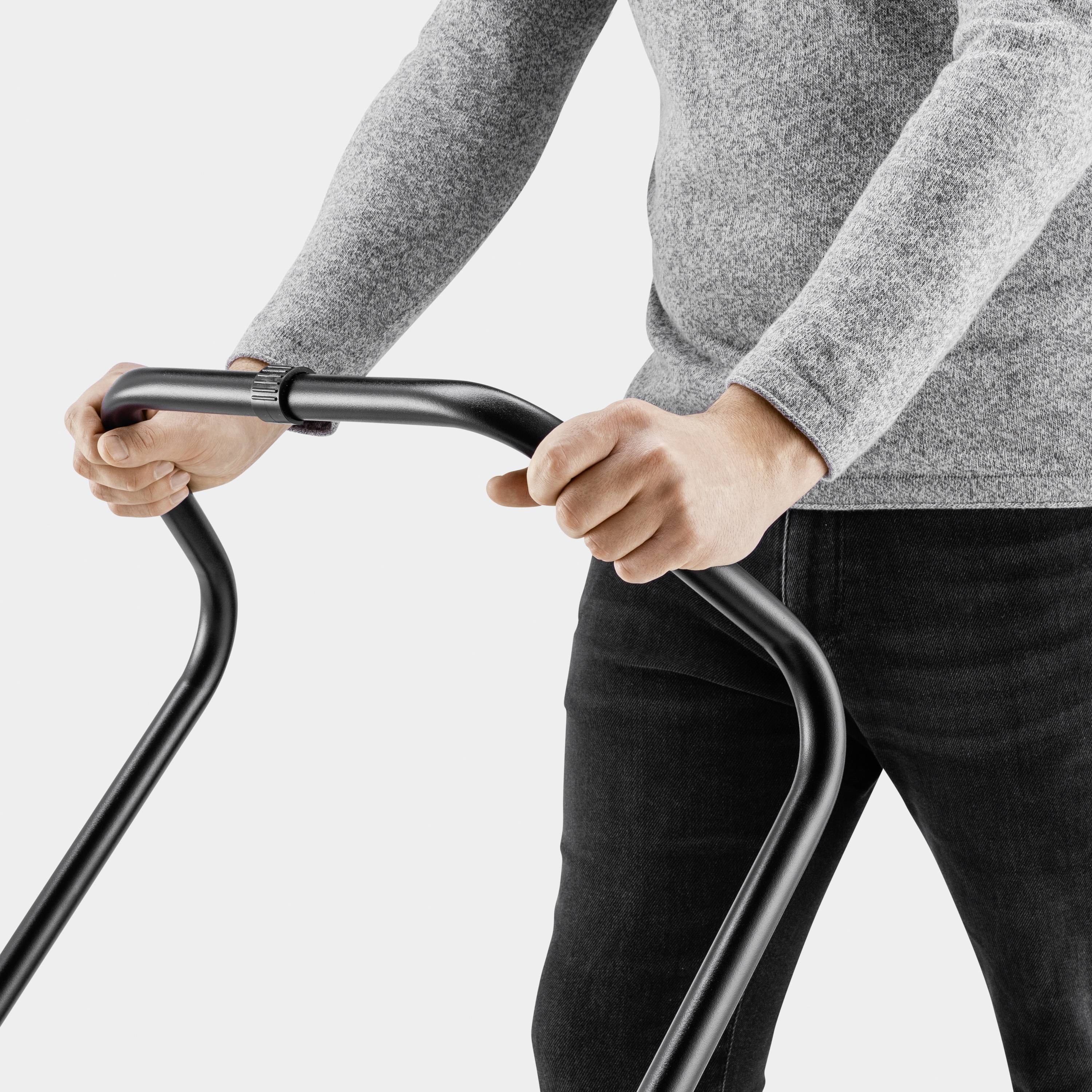 Une personne tient la poignée d'un déambulateur noir, porte un pull gris et un pantalon noir.