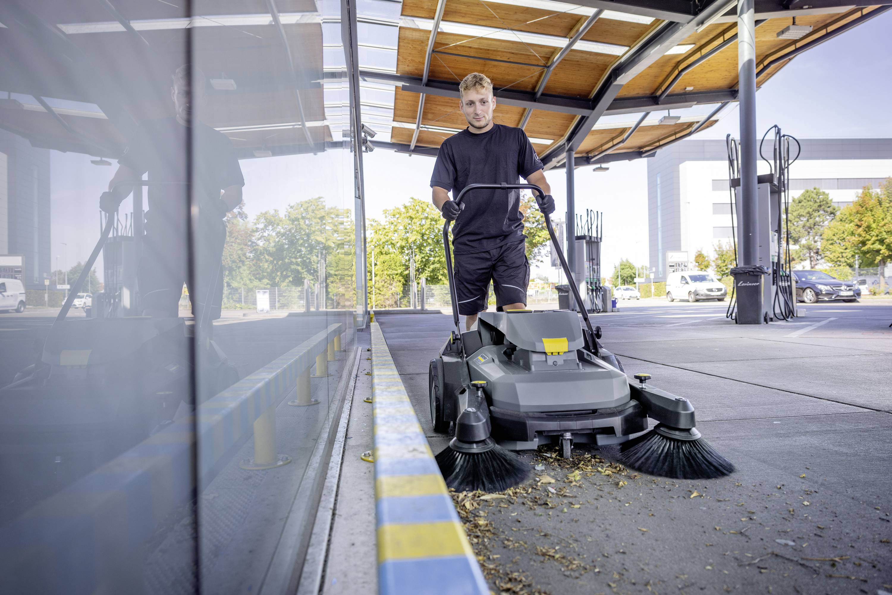 Un homme utilise un balai mécanique électrique dans un grand hall ouvert. En arrière-plan, on peut voir des arbres et des voitures garées.