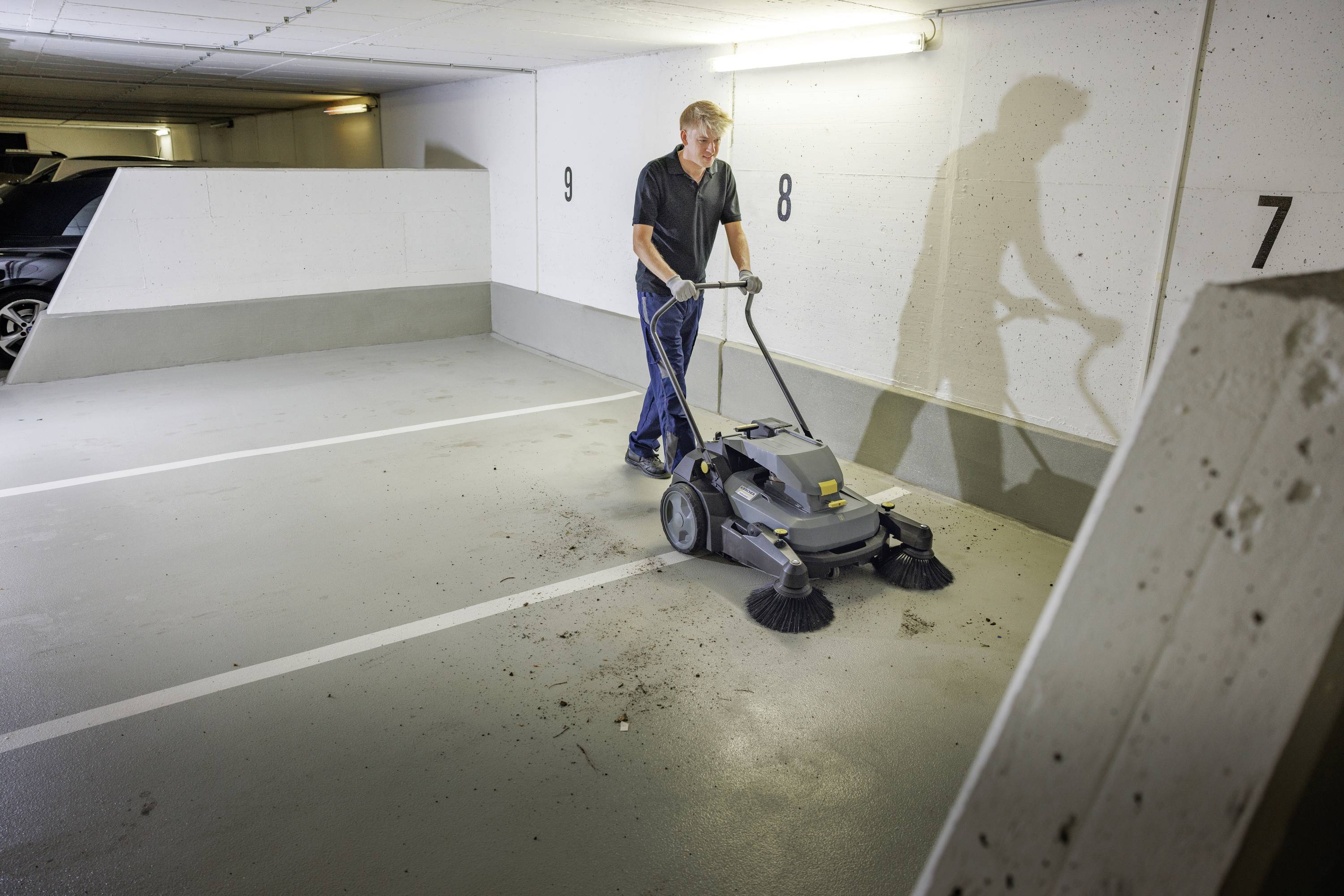 Une personne nettoie le sol d'un parking à l'aide d'une machine. La lumière venant de la gauche éclaire la scène. Des ombres sont visibles.