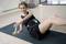 Une femme s'entraîne sur un tapis de yoga. Elle tient un petit haltère et porte un appareil de fitness autour de la taille. Elle sourit.