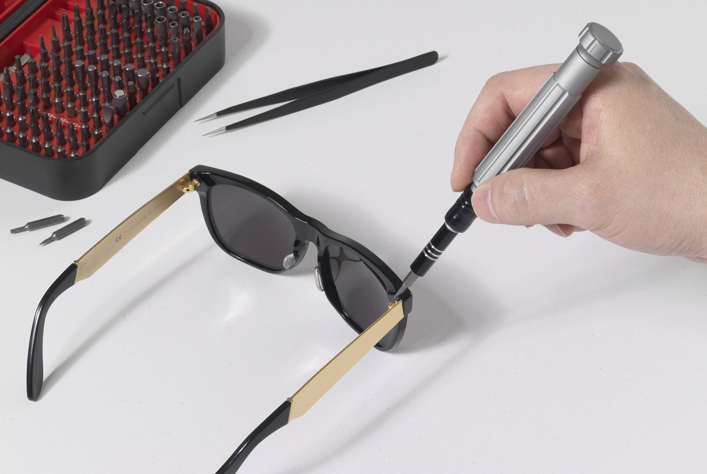 Une main répare une paire de lunettes de soleil avec un tournevis, entourée d'un ensemble d'outils variés.