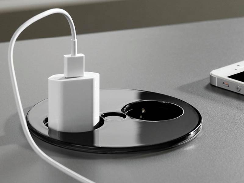 Un chargeur de smartphone branché dans une prise électrique intégrée à un bureau, avec un smartphone placé à proximité.