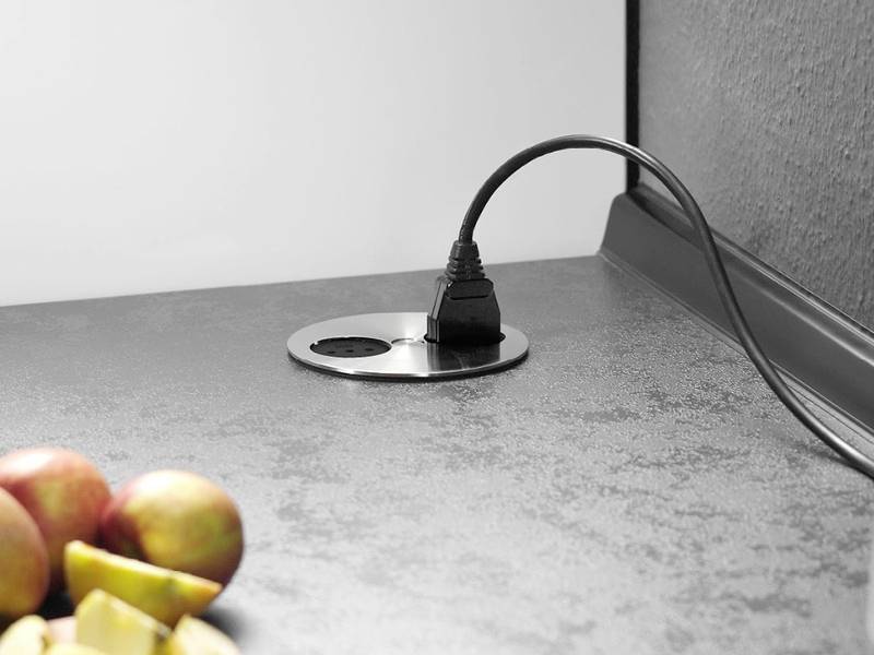 Un câble électrique branché dans une prise ronde sur un plan de travail de cuisine, avec des pommes et des tranches de pommes à proximité, indiquant un cadre domestique.