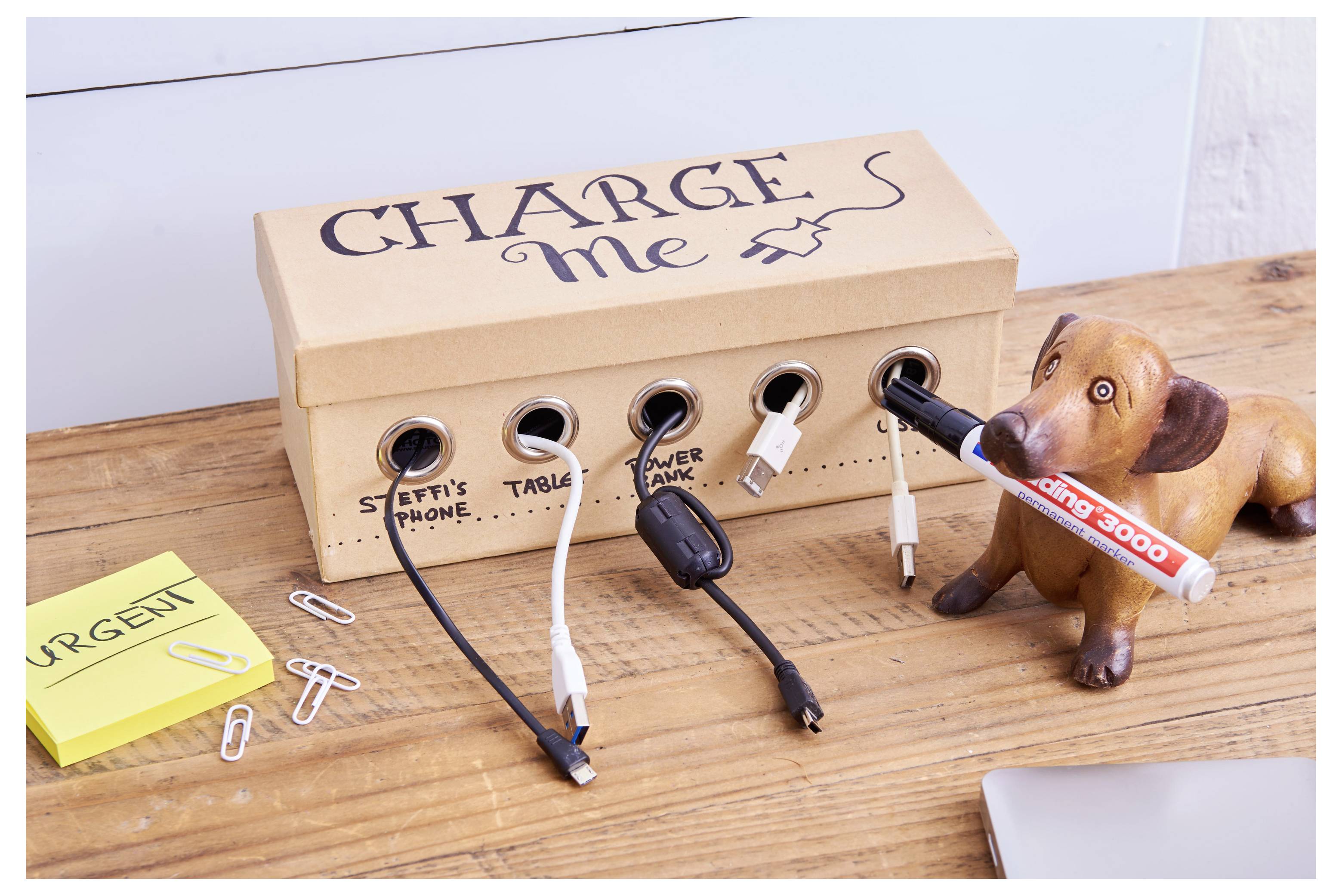 Station de recharge en carton humoristique intitulée « CHARGE me » avec plusieurs câbles d'appareils branchés, et un chien jouet tenant un stylo sur un bureau en bois.