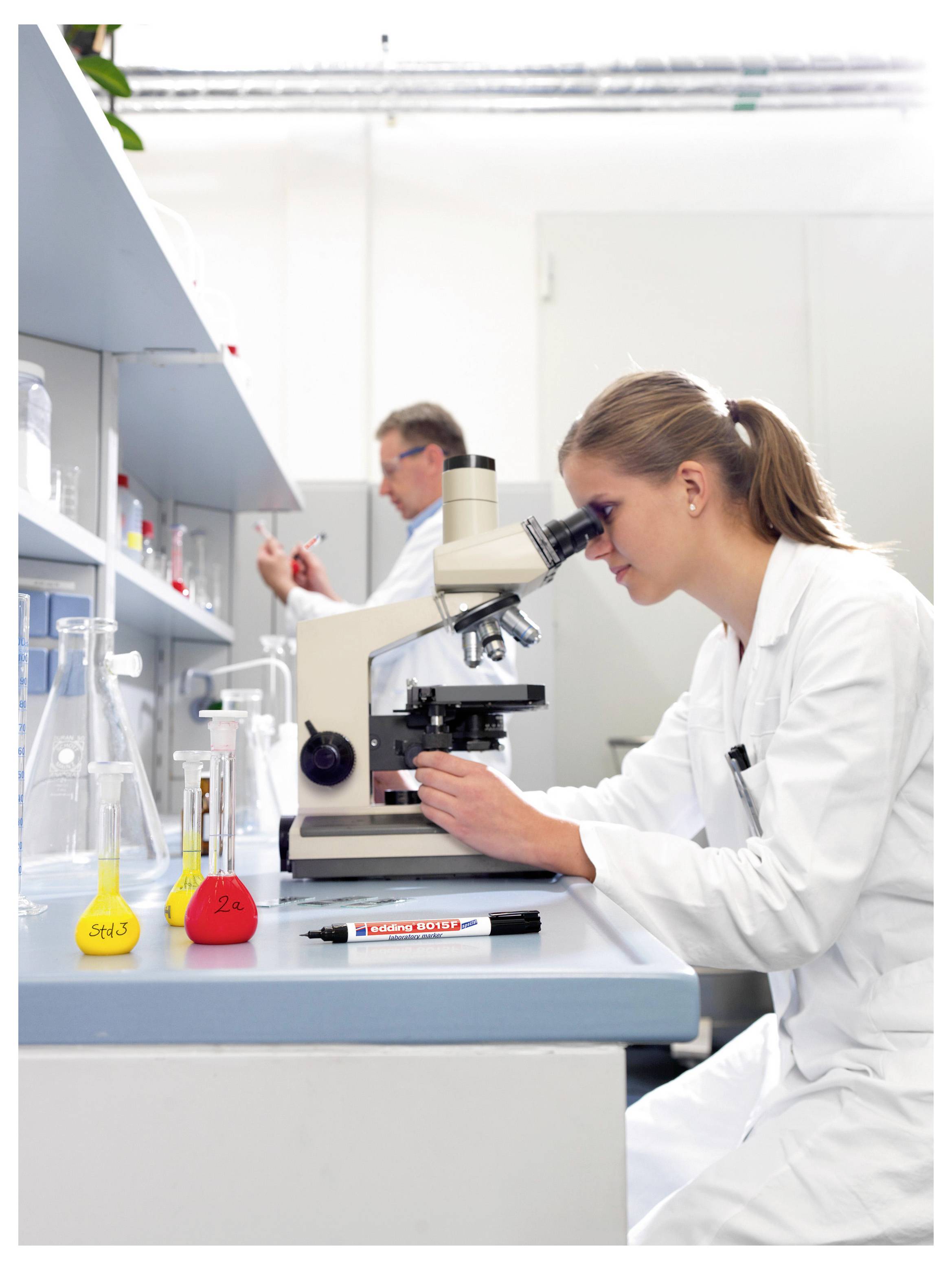 Scientifique en blouse de laboratoire regardant à travers un microscope sur un établi de laboratoire avec des fioles et du liquide coloré, tandis qu'un autre chercheur travaille en arrière-plan.
