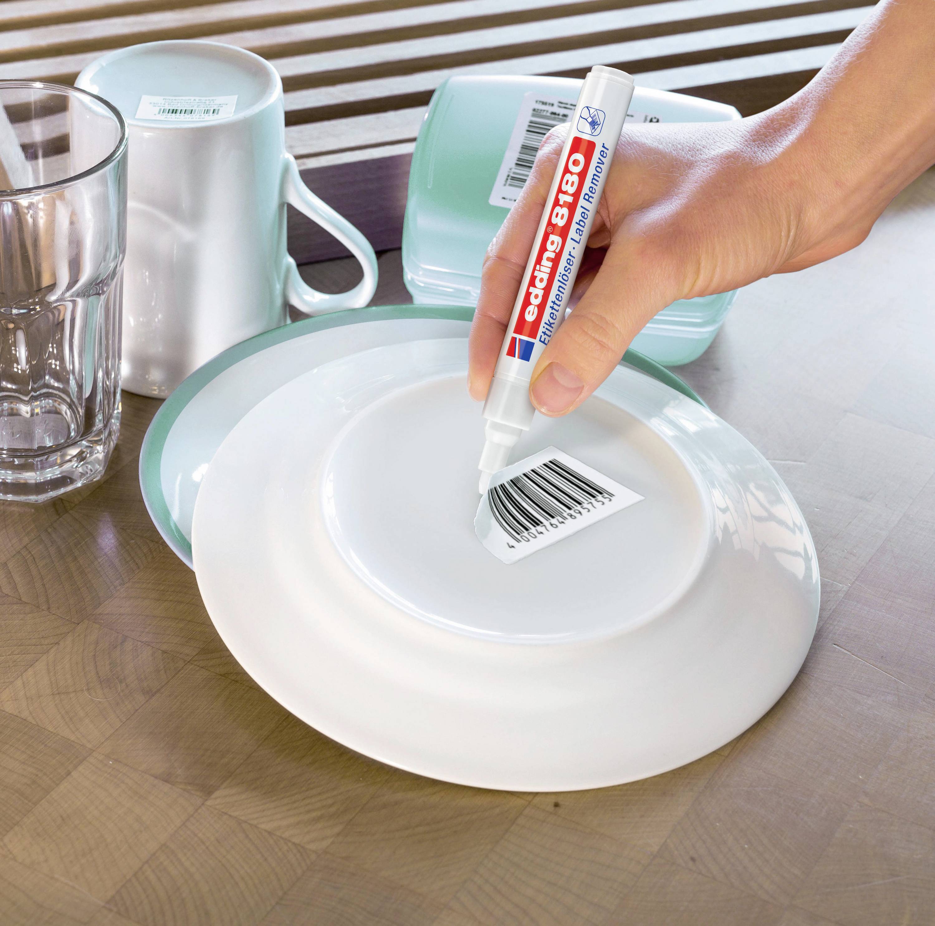 Une main tient un stylo spécial et efface un code-barres avec de la peinture blanche sur une assiette. En arrière-plan, une tasse et un verre sont posés.