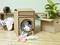 Un sèche-linge de jeu fabriqué en carton est posé sur un sol en bois. À côté, des vêtements colorés sont disposés et suspendus.