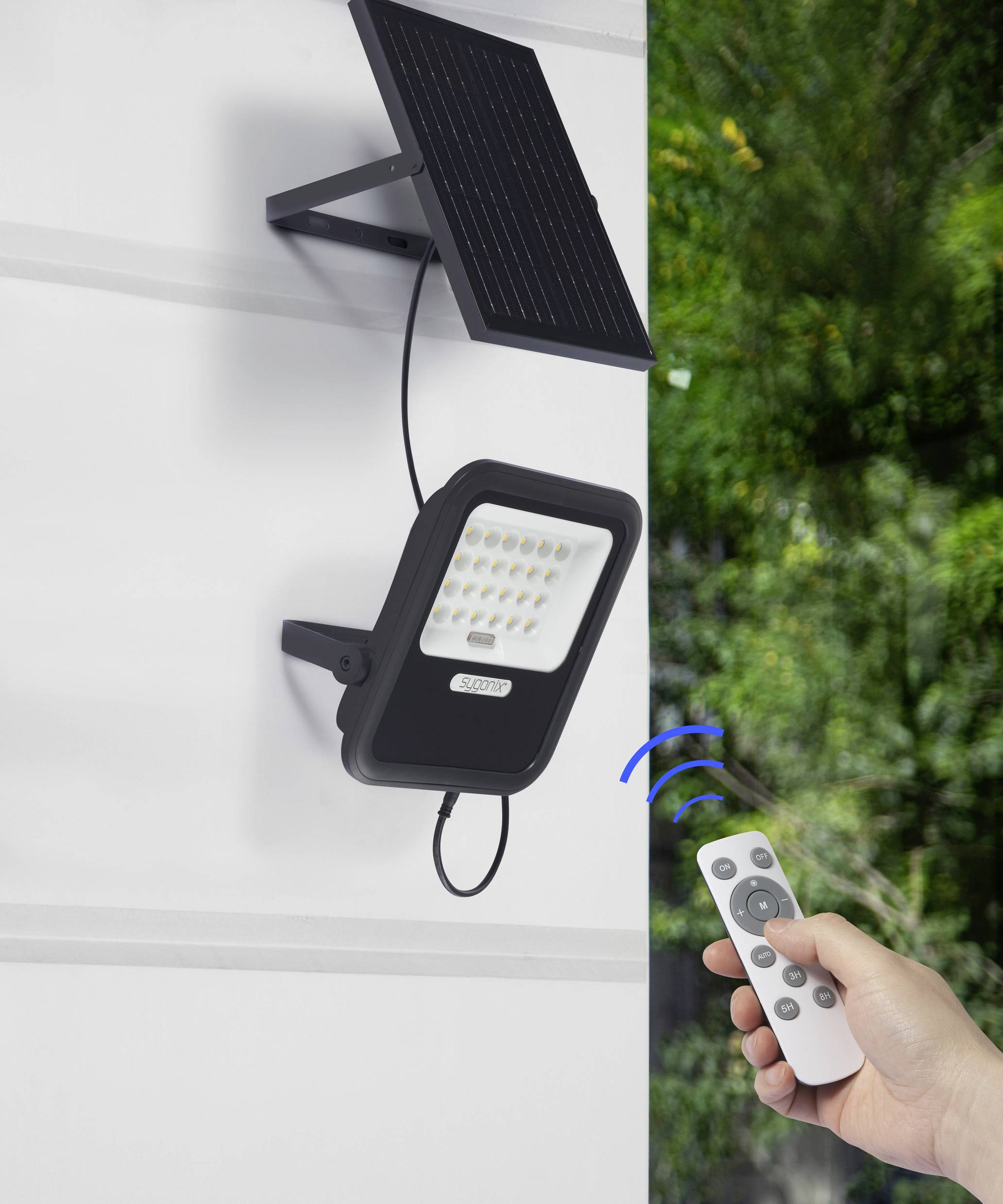 Lampe LED solaire montée sur un mur, contrôlée par une télécommande au premier plan. Arrière-plan : plantes vertes.