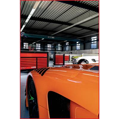 Dans un atelier, plusieurs voitures de sport et des armoires à outils rouges sont alignées. Au premier plan, on peut voir une voiture de sport orange.