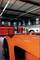 Dans un atelier, plusieurs voitures sont alignées, dont une voiture de sport orange au premier plan. À l'arrière-plan, on peut voir des armoires d'atelier rouges.